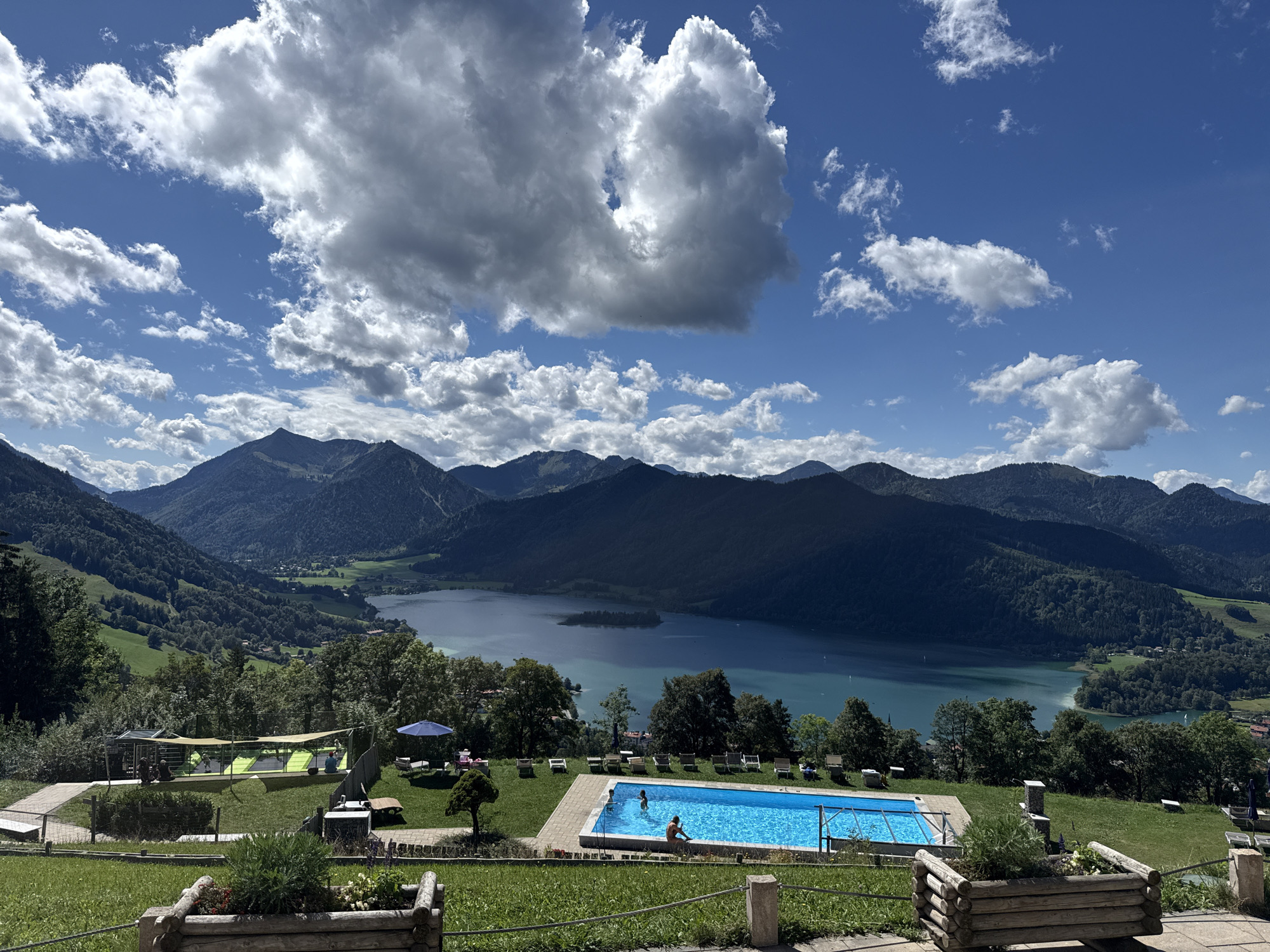 An aerial, wide-angle shot captures a scenic view of Schliersee, Germany, on a clear day with scattered clouds. In the foreground, a rectangular swimming pool with bright blue water is visible, with two people swimming in it. Surrounding the pool are lounge chairs and umbrellas, indicating a recreational area. Beyond the pool, a lush green lawn slopes down towards a lake, which is a prominent feature of the landscape. The lake is surrounded by rolling hills covered in dense green trees. In the background, a range of majestic mountains forms the horizon. The sky is a vibrant blue, with fluffy white clouds adding depth and texture to the scene. The overall impression is one of natural beauty and tranquility, with elements of human recreation integrated into the stunning natural environment.