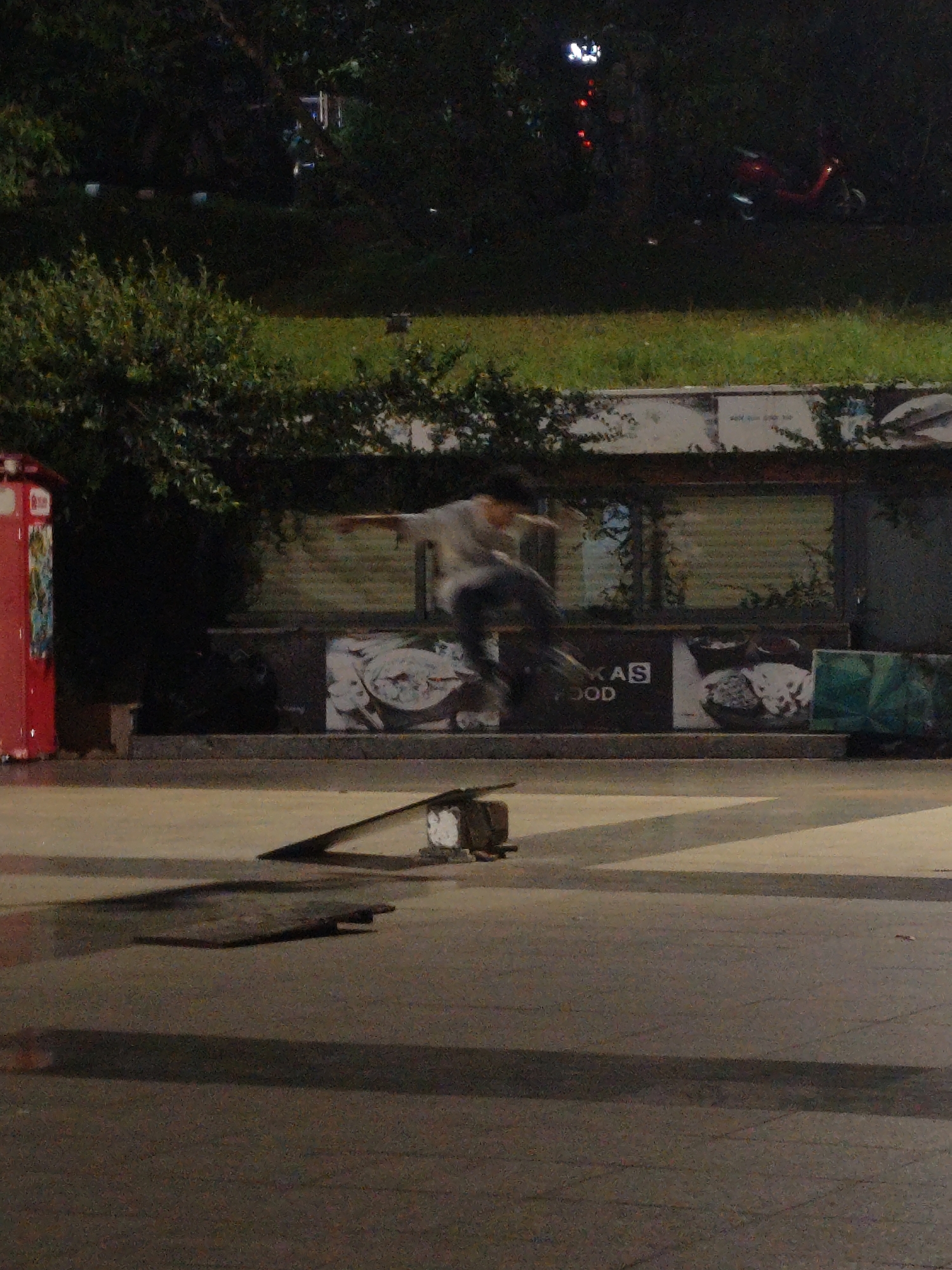 The image captures a dynamic street scene at night, likely in Da Lat, Vietnam, given the context. In the foreground, a skateboarder is captured mid-air, performing a trick over a ramp. The skateboarder, dressed in a light-colored shirt and dark pants, is blurred due to motion, indicating rapid movement. The ramp itself is a dark, angular object positioned on a paved surface. The background features a lush, green grassy area with trees. Further back, there are signs and a red structure that appears to be a kiosk or a public utility box. The overall atmosphere is dark, with artificial lighting creating highlights and shadows, suggesting an urban environment at night.