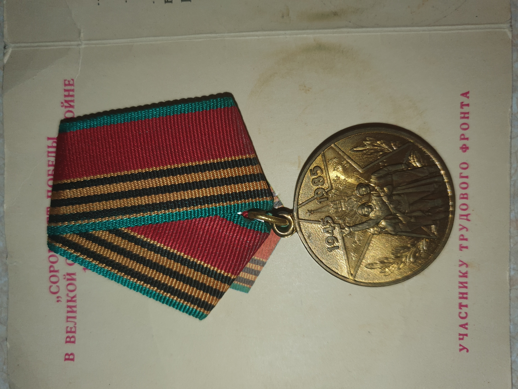 The image shows a close-up of a Soviet-era medal and its accompanying documentation. The medal is circular and made of a metallic material, possibly bronze, with a raised relief design. It features the years "1945" and "1985" on the upper portion, with a star and imagery of soldiers or workers. The medal is suspended from a ribbon with alternating stripes of red, black, and green. The documentation is a light-colored card or paper with text printed in Russian. The visible text on the documentation includes phrases like "ВЕЛИКОЙ ПОБЕДЫ" (Great Victory) and "УЧАСТНИКУ ТРУДОВОГО ФРОНТА" (To the participant of the labor front). The overall scene is static and appears to be a still life of a historical artifact. There is no indication of the city of Tashkent, Uzbekistan, or any specific location within it. The lighting is even, and the focus is on the medal and the text.