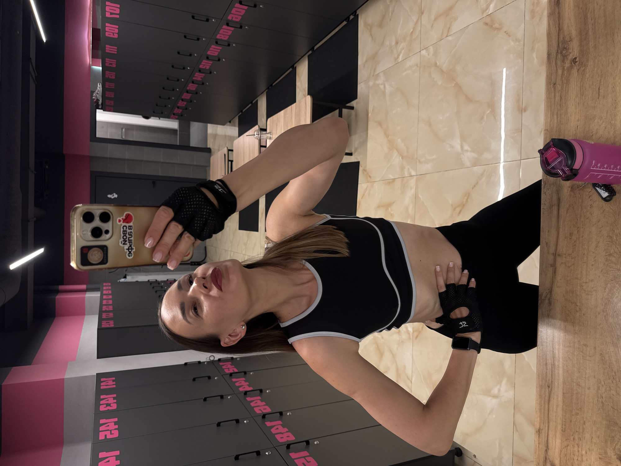 A young woman with brown hair is taking a selfie in a gym locker room. She is wearing a black sports bra with a grey trim and black leggings. She has on black fingerless gloves and a black fitness tracker on her left wrist. The locker room has dark grey lockers with pink lettering and numbers. The floor is tiled with a marble pattern. To the right of the woman, a pink water bottle with measurement markings is visible. The woman is holding her phone in her right hand, which displays a sticker of a cartoon character and the text "B SUPO CBON" and "IOS 108 111 113 120". The overall impression is of a workout in progress, with the woman likely in between exercises or having just finished. The lighting suggests it is daytime. The image is oriented vertically.