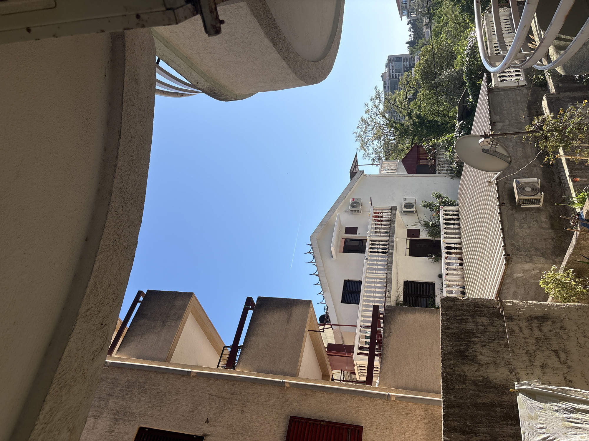 The photograph depicts an upward-angled view of buildings in Bečići, Montenegro, seen from a courtyard. The perspective is framed by parts of buildings on either side, creating a narrow view toward a clear blue sky.
The building directly in the center of the frame is white with multiple stories, visible air conditioning units, and decorative railings. A metal staircase is visible on one side of this building.
To the right, there is a building with a corrugated metal roof, satellite dish, and an air conditioning unit. The building to the left is beige with a distinct, angled roof.
The overall scene is captured in sunlight, with details in the architectural structures and their textures. The image captures a moment within the dense building environment of the Bečići urban landscape.