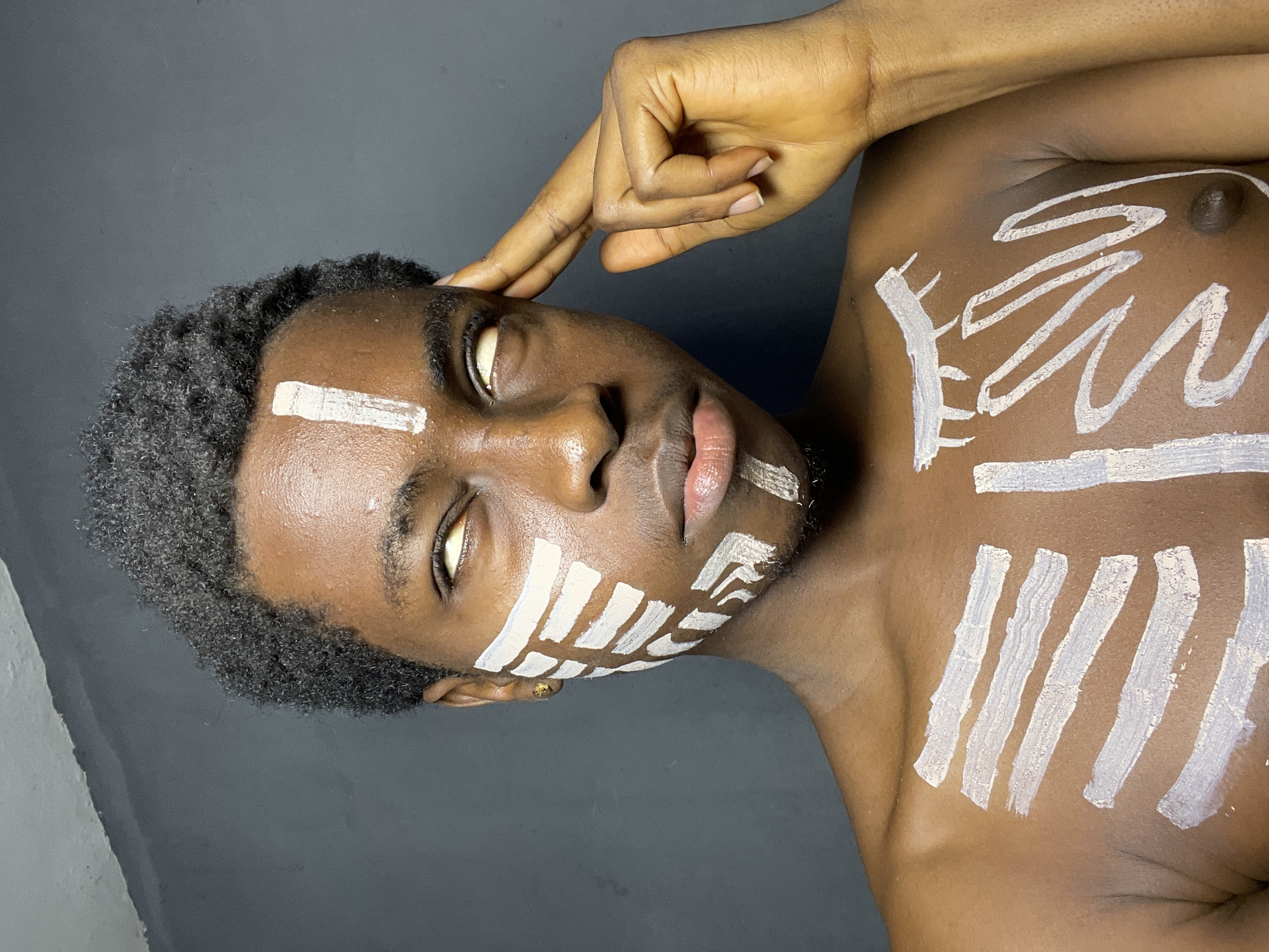 A medium shot depicts a Black man with close-cropped, curly black hair, wearing white paint markings on his face and upper chest. He is positioned horizontally with his head tilted slightly upwards. The paint on his face forms geometric shapes around his eyes, cheeks, and forehead. The markings on his chest appear as a combination of straight lines and wavy shapes. His eyes are partially closed, and his skin appears oily. His right hand is raised and positioned near his forehead with fingers lightly touching it. The background is a solid, dark gray color. No indication of the city of Nkpor, Nigeria is evident in the image.