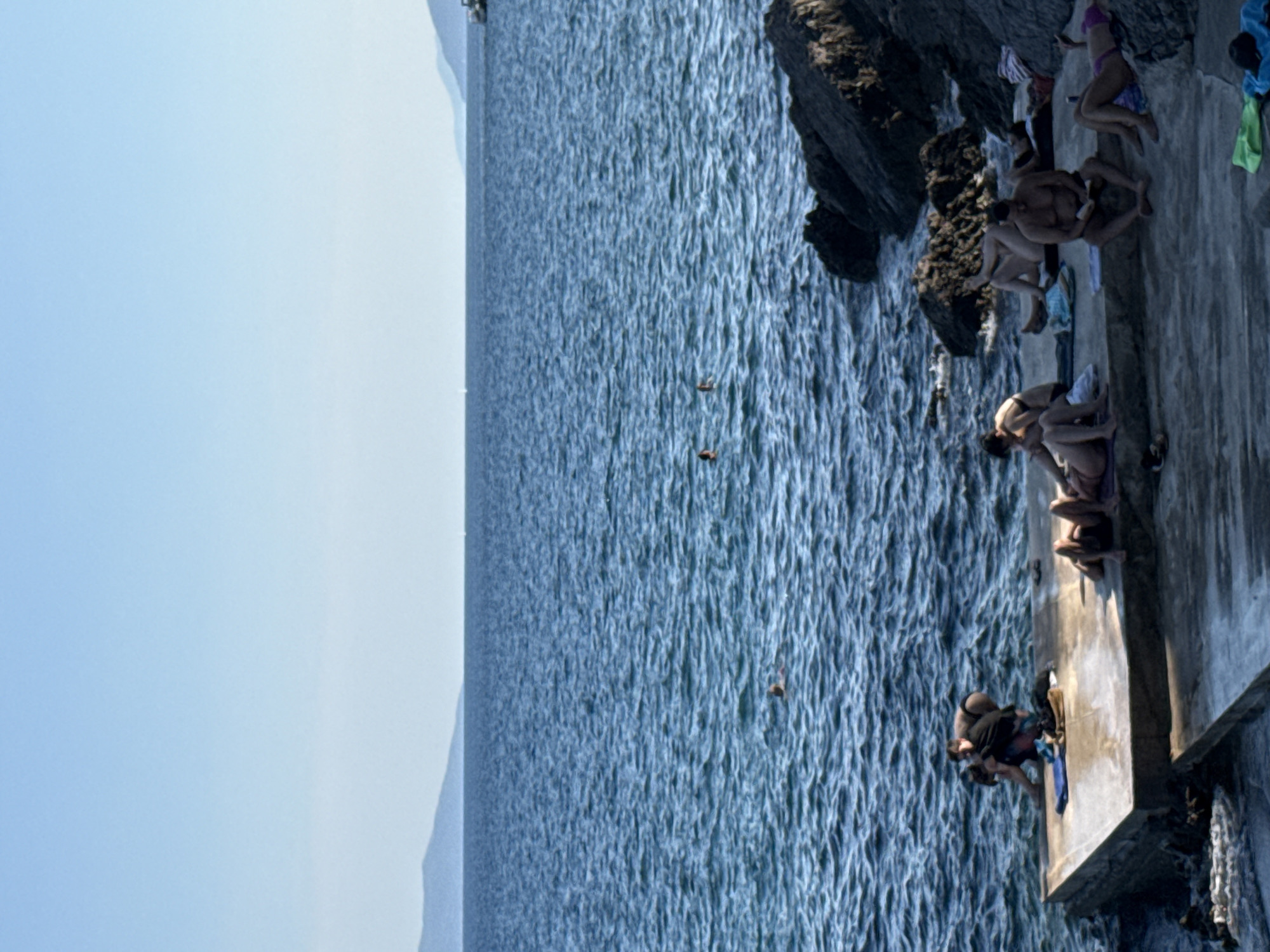 In Grad Opatija, Croatia, on a sunny day, several people are relaxing on a concrete platform adjacent to a rocky shore. The sea is a deep blue with gentle waves. In the foreground, on the platform, a group of individuals are sunbathing. One man sits with a book, while others recline on towels. Towards the left, a couple is engaged in an interaction near the water's edge. Further out in the sea, a few swimmers are visible. The background features a clear sky and distant hills or mountains.