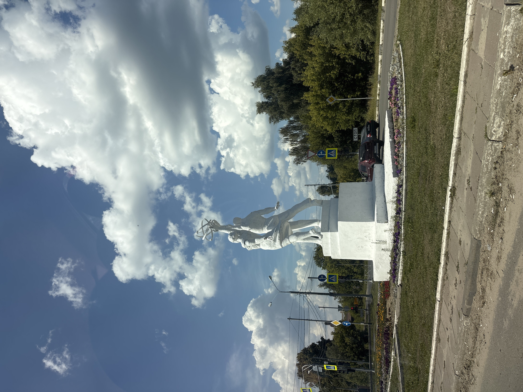 This media file captures a low-angle, vertical shot of a white stone sculpture depicting two figures reaching upwards, holding a spherical object with protrusions. The sculpture stands on a white pedestal in what appears to be a public space. Behind the sculpture, a dense line of green trees forms a backdrop, with a clear blue sky dotted with large, fluffy white clouds above.
In the foreground and mid-ground, the scene includes elements typical of a street or park area. There are several road signs visible, including a yellow diamond-shaped sign and a blue circular sign with an arrow. A yellow pedestrian crossing sign is also present, indicating a road or intersection nearby. Two cars are parked along the side of a road, one dark red and one darker colored. A patch of green grass, with a curved border of small purple and yellow flowers, runs alongside a paved pathway and a road. The weather appears to be sunny with scattered clouds, suggesting it is daytime. The overall mood is peaceful and static, with the focus on the monument and the natural surroundings.
Visible text includes the word "СТОП" (STOP in Russian) on a road sign. The location context is identified as Zavolen'e, Russia.