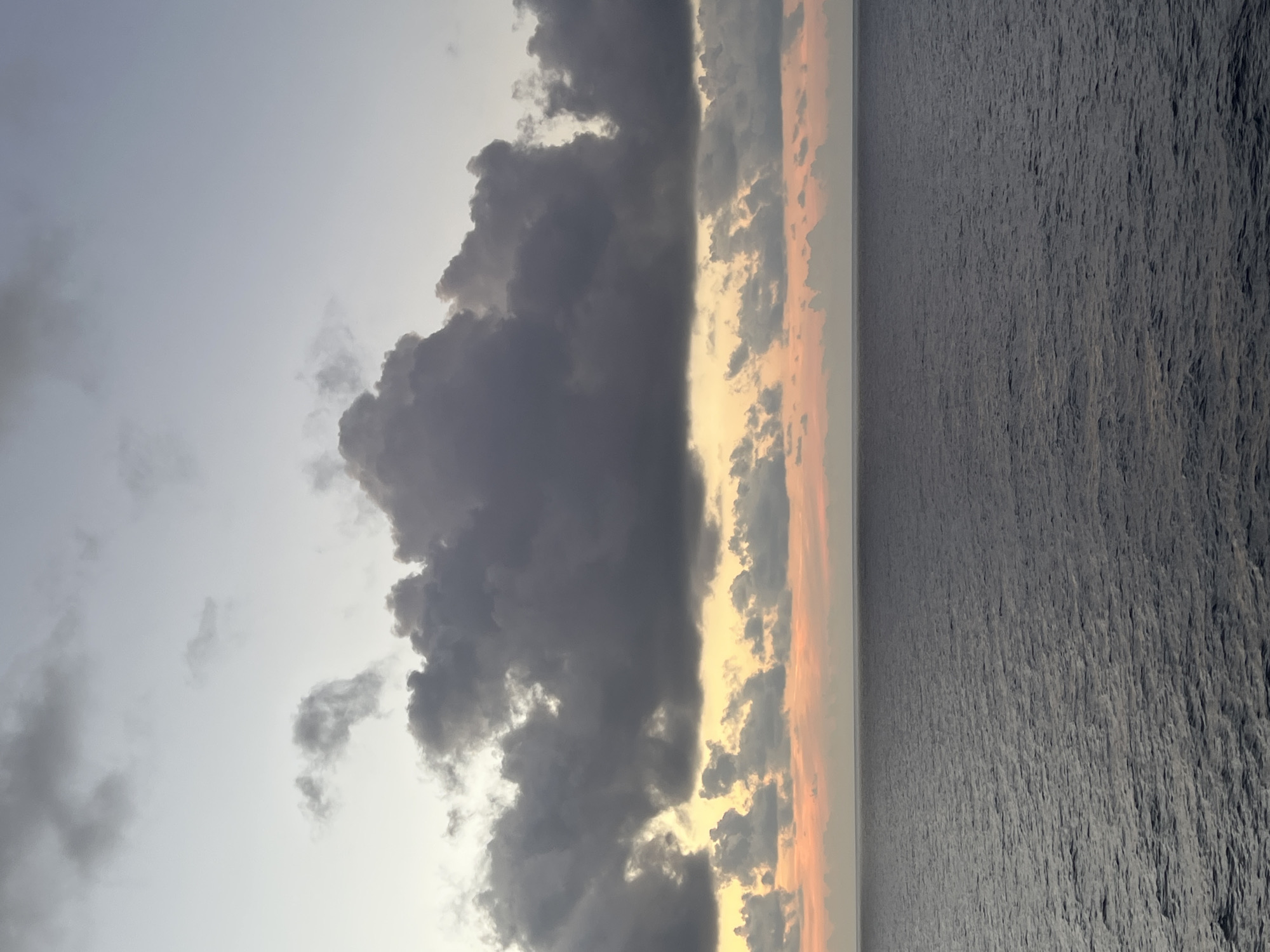A wide shot captures a dramatic sky over the ocean. Dark, heavy clouds dominate the upper portion of the image, with lighter, wispy clouds and streaks of orange and pink hues present lower in the sky, suggesting a sunrise or sunset. The horizon line is sharp, dividing the sky from the vast, dark blue ocean which fills the bottom half of the frame. The water appears choppy with small waves and ripples. The scene is devoid of any man-made structures or living beings.