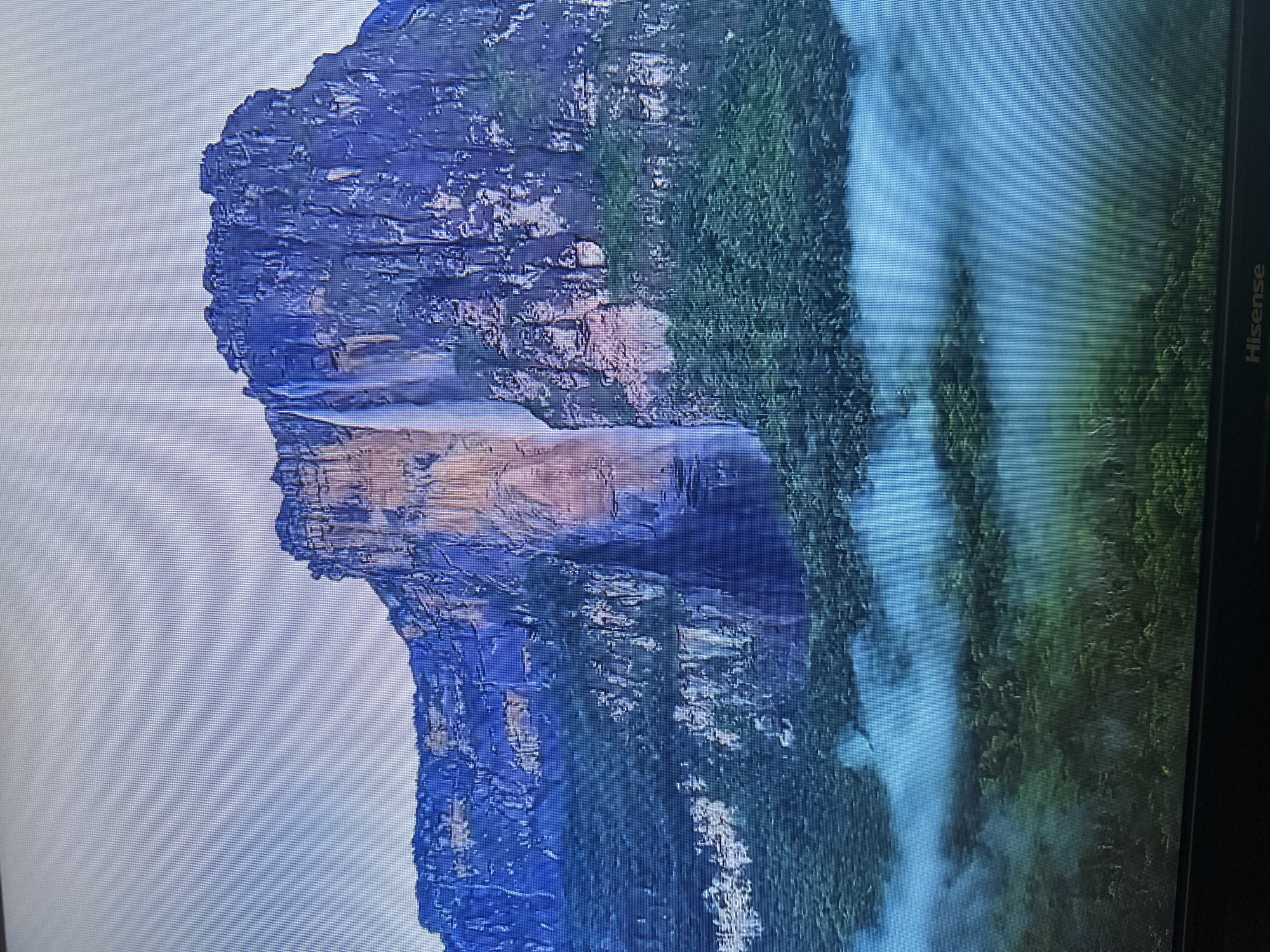 The image shows a wide shot of Angel Falls, the world's tallest uninterrupted waterfall. The waterfall cascades down a sheer cliff face, which is covered in lush green vegetation at its base. A dense jungle can be seen below the waterfall, partially obscured by a thick layer of blue mist or clouds. The sky above is a pale, overcast blue. The scene is devoid of any human presence or structures, suggesting a remote, natural environment. The image is not located in the city of Kyiv, Ukraine, as it depicts a natural landscape in Venezuela.