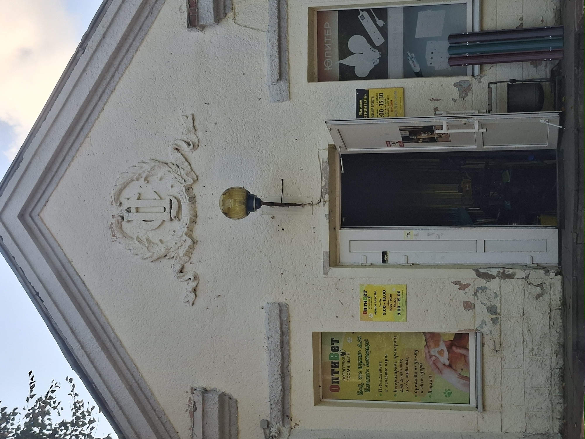 The image shows the exterior of a building with a light-colored facade. The building has a peaked roof with decorative trim and a large, ornate relief above the entrance. There is a vintage-style yellow globe lamp attached to the wall to the right of the entrance.
The entrance is a double door, with the left door ajar, revealing darkness within. To the left of the entrance is a window displaying information for "Оптивет" (OptiVet), a veterinary clinic and pet store. The poster lists services such as daily and therapeutic food, veterinary preparations, grooming supplies, and vaccines. Below this poster is another smaller sign for "Оптивет" detailing their work hours.
To the right of the entrance is a window with the word "Юпитер" (Jupiter) displayed, and images of light bulbs and an electrical extension cord, suggesting it is an electrical or hardware store. Below this window, leaning against the building, are three vertical metallic poles in burgundy, green, and dark grey.
A trash can is located to the right of the open door. The building's facade shows signs of wear, with peeling paint and chipped plaster. The ground in front of the building is paved. The sky is visible in the background with some clouds, and a tree branch with leaves is seen in the upper left corner. The overall impression is that of a commercial building in a somewhat weathered state.