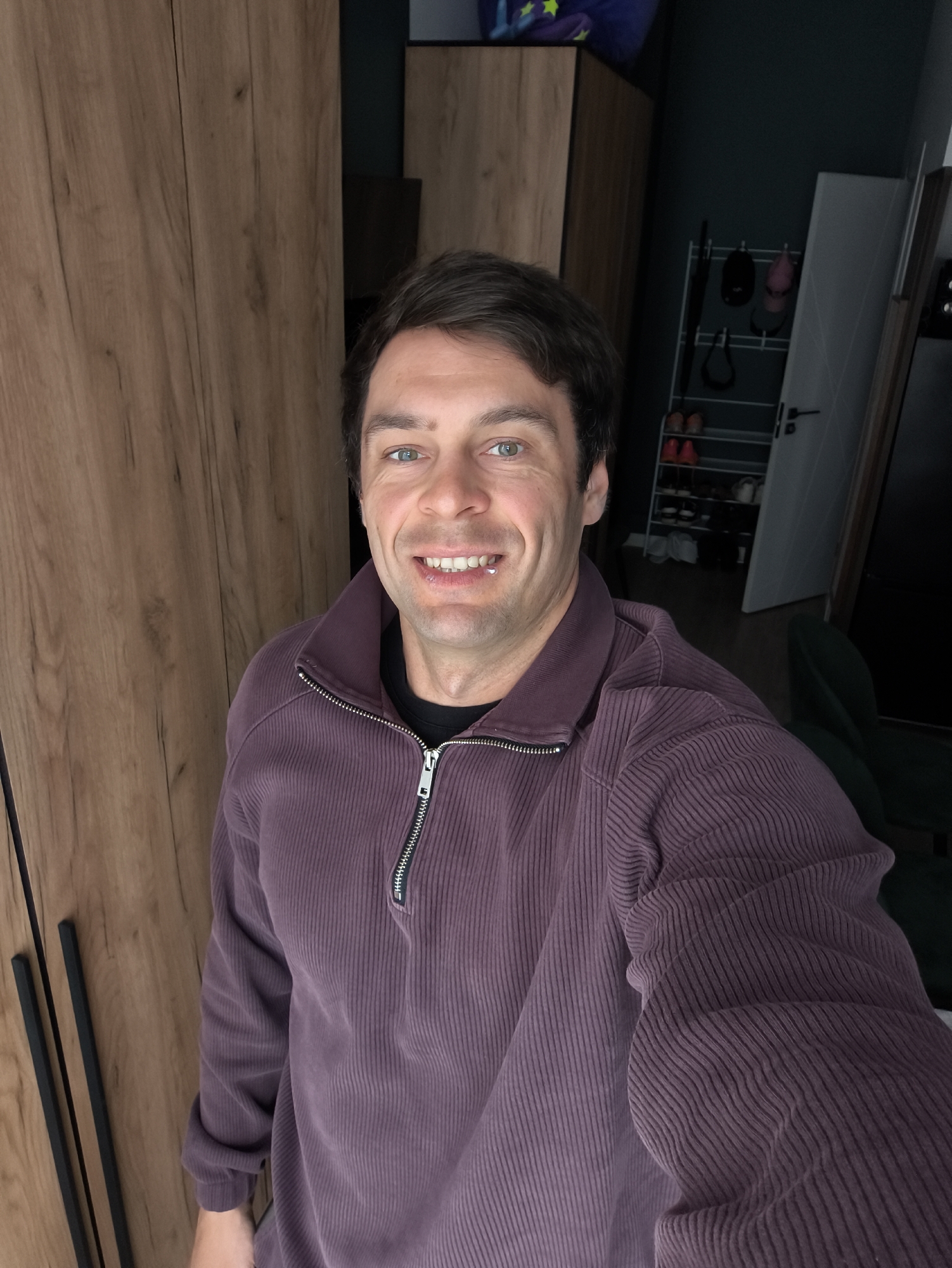 A man is taking a selfie indoors. He has short dark hair, blue eyes, and is wearing a purple corduroy zip-up shirt over a black t-shirt. Behind him are wooden wardrobes and a doorway leading to a dimly lit room. In the background is a white shoe rack with shoes, hats, and an umbrella. There's a green upholstered chair partially visible in the darker room. The location appears to be inside a residence. There's no information to indicate this is Moscow, Russia.