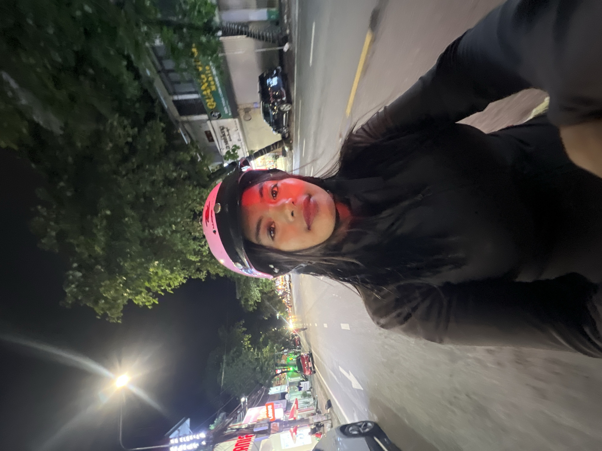 The image is a selfie taken at night from the perspective of someone riding a motorcycle. The subject is a woman wearing a pink helmet and a black jacket. She is looking directly at the camera with a neutral expression. The background shows a street scene with buildings, trees, streetlights, and other vehicles. The street appears to be well-lit, indicating an urban environment. The presence of businesses with signage suggests this is likely a commercial area within Minh Xuân Ward, Vietnam, possibly near a main road given the traffic. The overall atmosphere is that of a typical city night, with the artificial lights illuminating the scene.