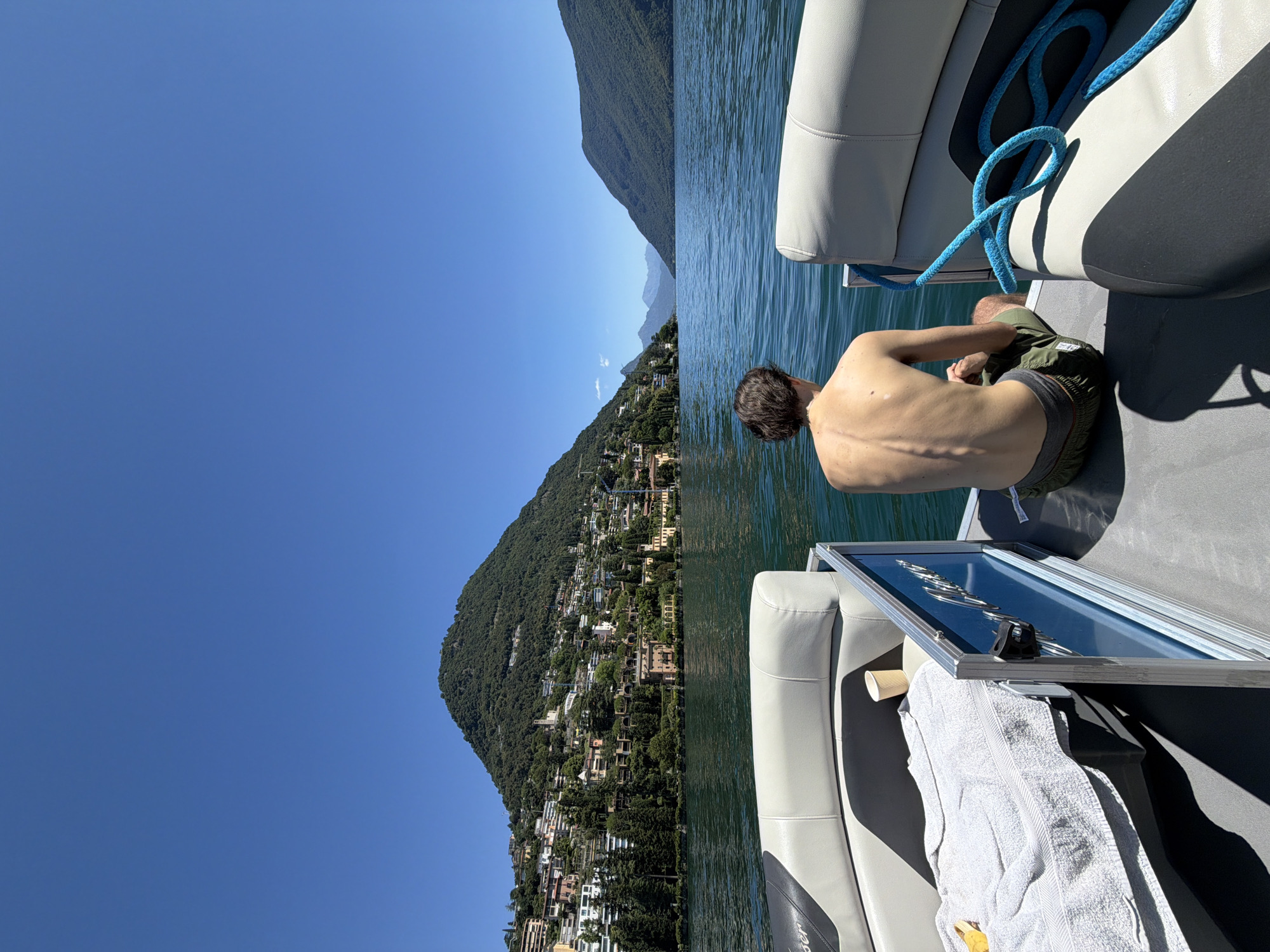 In Lugano, Switzerland, a young person is seated on the rear deck of a boat, facing away from the camera. The person is shirtless, wearing green shorts, and their back is visible, showing a lean physique. The boat, a pontoon style, has white and grey seating and a blue railing with a boat nameplate. A white towel and a paper cup are on the deck to the left of the person. A bright blue rope is coiled on the right side of the boat. The boat is on a body of water, likely Lake Lugano, with a lush green mountain covered in buildings and trees dominating the background on the left side. The sky is clear and blue. To the right, the lake extends towards more mountains in the distance. The scene captures a moment of leisure on the water on a sunny day.