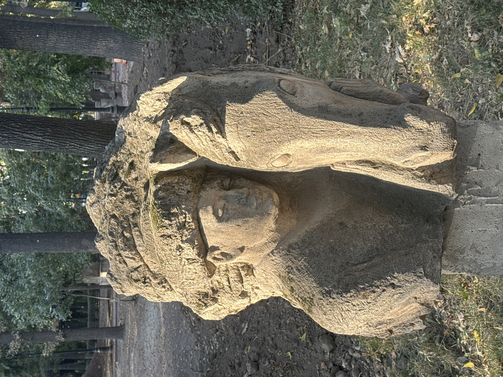 A stone sculpture of a man and a horse is centrally located in a park in Bishkek, Kyrgyzstan. The sculpture, carved from a light-colored rough stone, depicts a man's head and upper torso nestled against the head of a horse. The man appears to be wearing a hat. The horse's head is positioned to the right of the man's, with its eyes and muzzle clearly defined. The sculpture is set on a stone pedestal and is surrounded by trees, grass, and dirt ground. The lighting suggests it is daytime, with sunlight casting shadows on the sculpture and the surrounding area. In the background, more trees and park elements like lamp posts are visible.