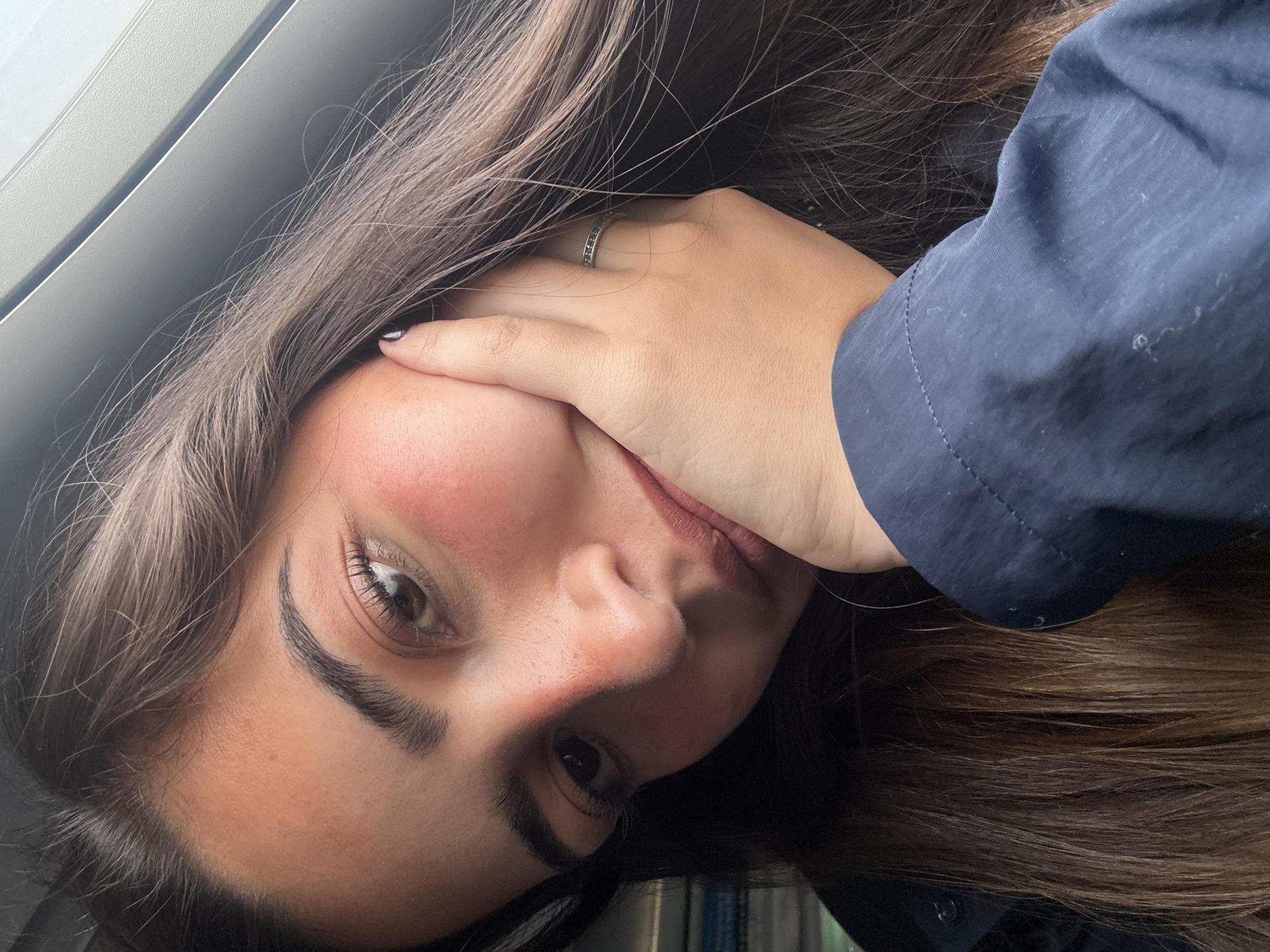 A young adult female is pictured in a close-up shot. She has long brown hair and is wearing a blue button-up shirt. Her left hand is partially covering her face. The photo was taken indoors, with a window visible in the background. The location cannot be determined as San Antonio, United States of America based on the image provided.
