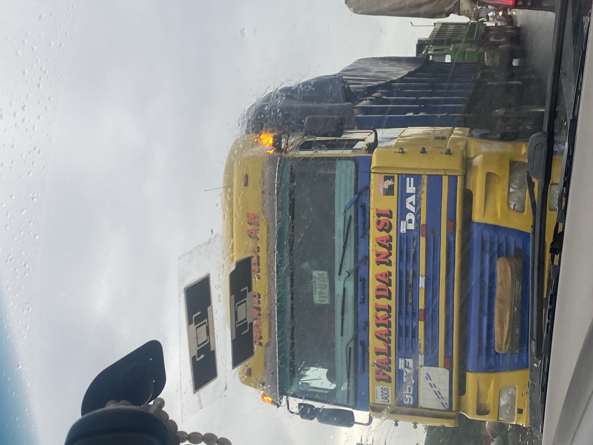 The image shows a yellow and blue DAF truck, likely in Keffi, Nigeria, with rain visible on the windshield, obscuring some details. The truck has "FALAKI DA NASI" written in red and yellow lettering on its side, along with "LAGOS" and "95 XF". The truck is in traffic, as other vehicles are visible behind it. The sky is overcast, suggesting a cloudy or rainy day. The scene is a street view, likely from another vehicle, looking forward at the truck.