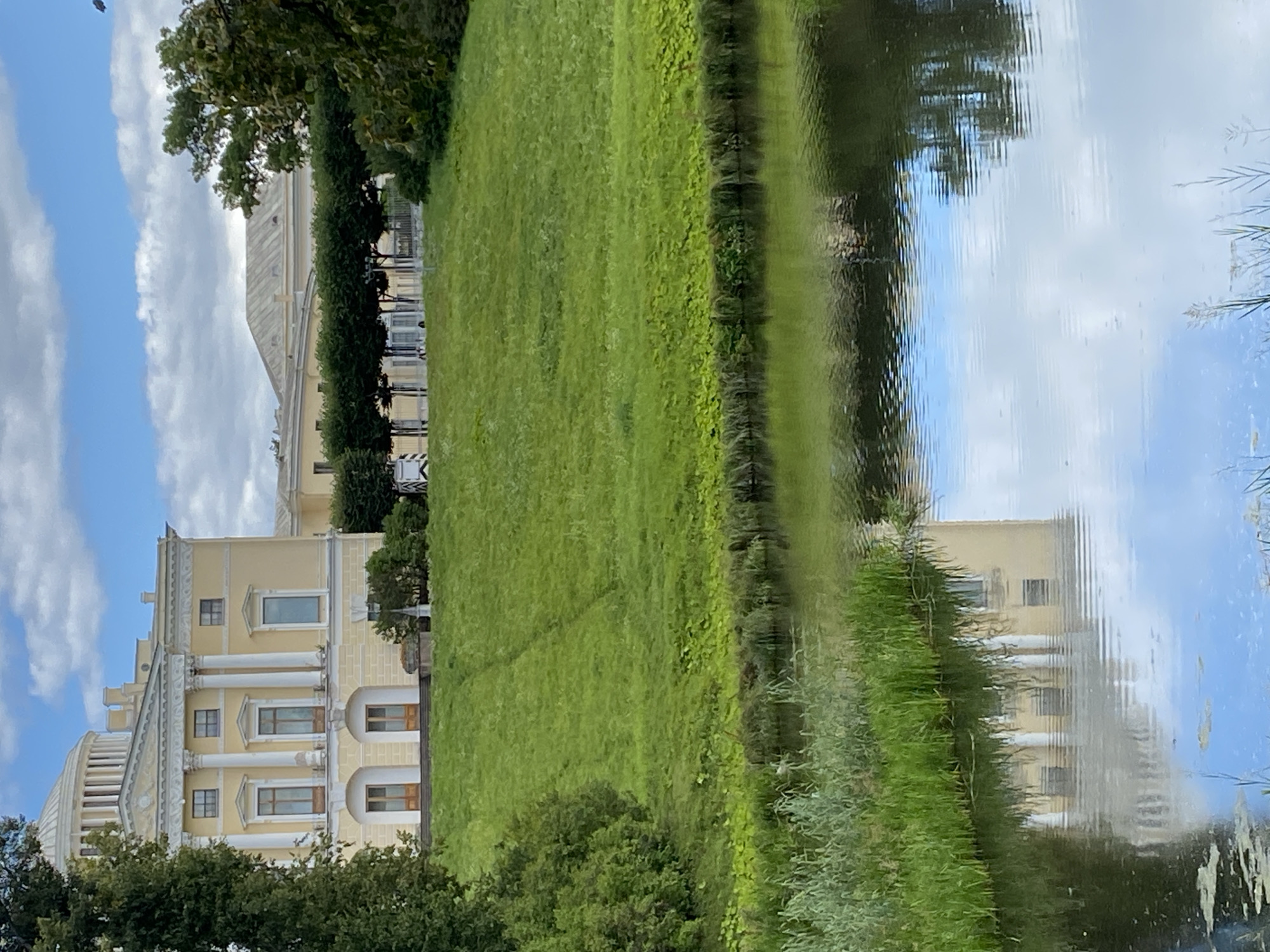 The image captures a view of the Pavlovsk Palace in Pavlovsk, Russia. The palace, painted in a soft yellow hue, features classical architecture with prominent columns and a domed roof. It is situated atop a gently sloping grassy hill. In the foreground, a calm body of water reflects the sky, clouds, and the palace itself, creating a serene atmosphere. Lush green foliage and tall trees frame the scene, adding to the natural beauty of the park setting. The sky is partly cloudy with white cumulus clouds against a blue background.