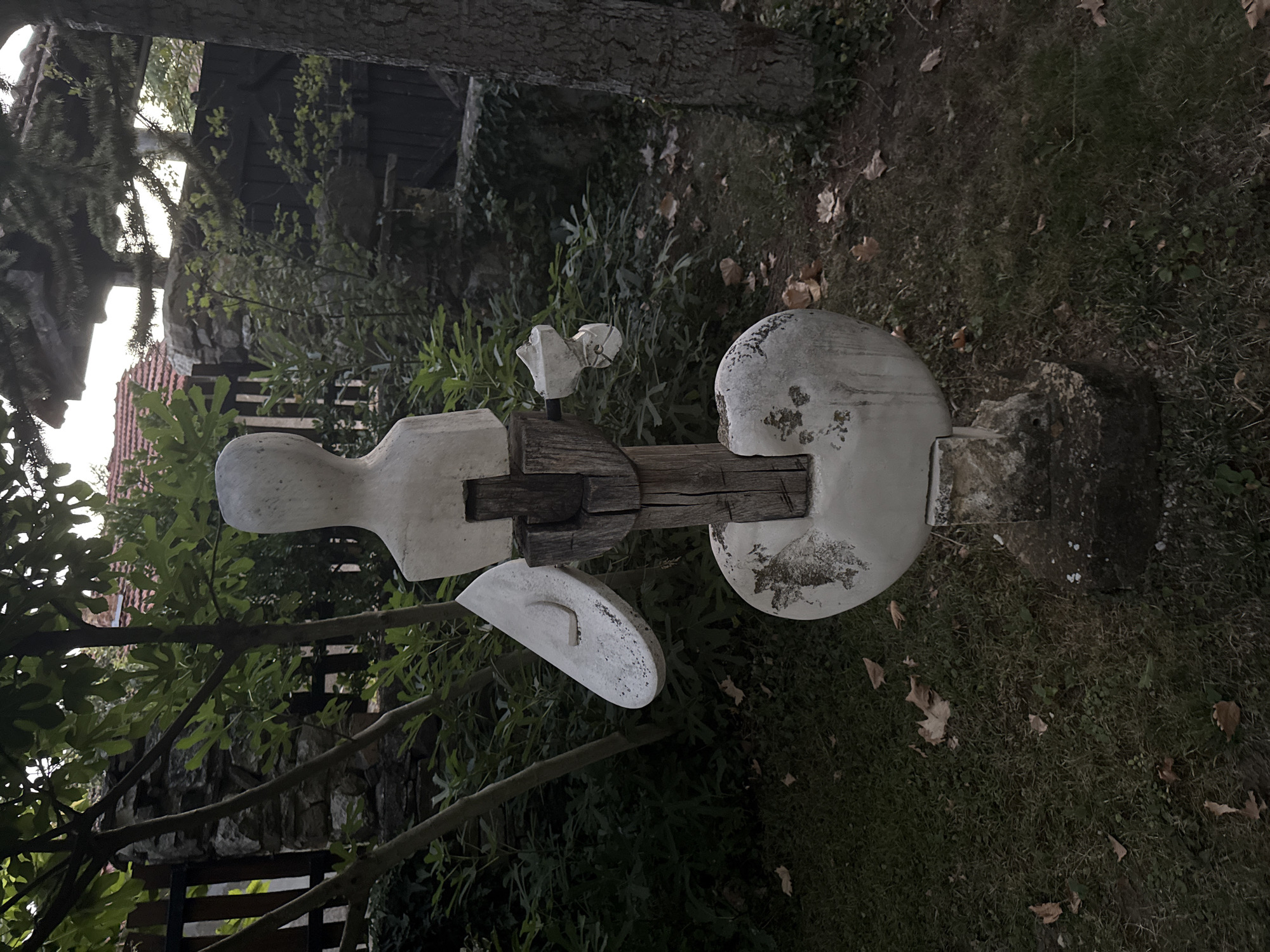 The image depicts an abstract sculpture made of concrete and wood, standing in a garden. The sculpture is positioned centrally in the frame. It features a headless torso-like form, a large, flattened oval shape at the base, and two disc-like elements attached to the sides of the torso. Wooden beams are integrated into the structure, particularly for the torso. The sculpture is weathered and shows signs of age.
The surrounding environment is a garden with lush green foliage and trees. Fallen leaves are scattered on the grass. In the background, there are hints of a fence made of stone and wood, and parts of buildings with red tile roofs, suggesting a residential area. The lighting suggests it is daytime, possibly late afternoon, with diffused light filtering through the trees. The location is identified as Petrevene, Bulgaria. There are no people or animals visible in the image.
