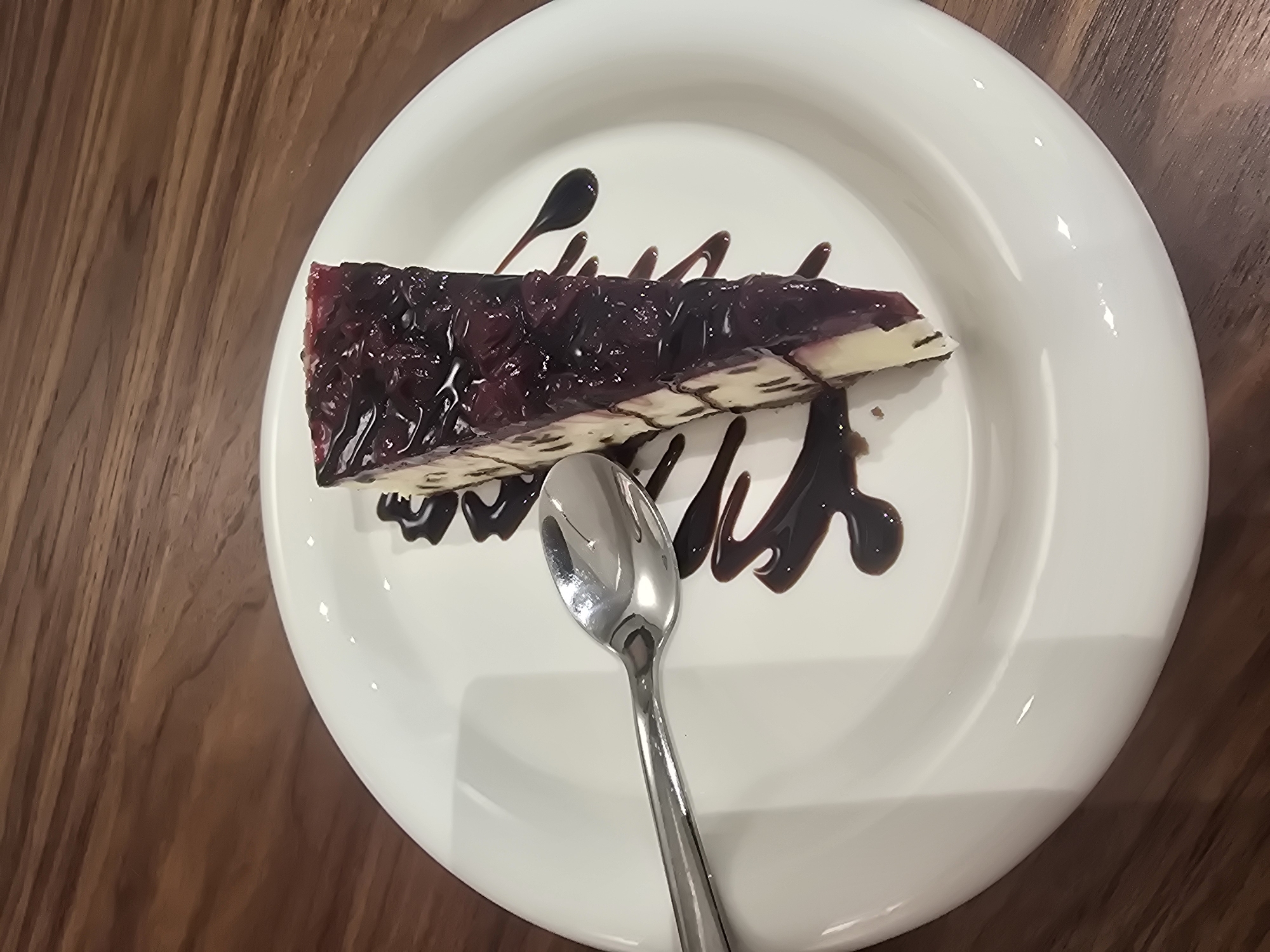 A slice of blueberry cheesecake sits on a white plate, adorned with a drizzle of chocolate sauce. A silver spoon rests on the plate to the left of the cheesecake. The plate is positioned on a wooden surface, likely a table. The background is blurred, but the texture of the wood is visible. The scene appears to be indoors, in a restaurant or cafe setting. There are no people visible in the image. The lighting is natural or ambient, creating soft shadows. There is no information in the image to suggest it is located in Ulan-Ude, Russia.