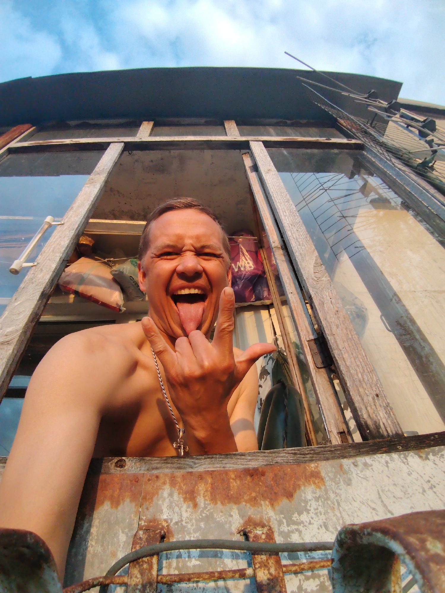 A low-angle, wide shot shows a shirtless man sticking his tongue out and making a rock-and-roll hand gesture from a window. The man, who appears to be in his 40s, has short brown hair and is wearing a silver chain with a cross pendant. He is framed by a weathered wooden window frame. In the background, various objects are visible, including bags, a ladder, and what appears to be an antenna. The sky above is blue with some clouds. The overall scene suggests an urban environment, possibly in Chaikovsky, Russia, given the context. The image is taken from a street-level perspective, looking up at the man in the window.