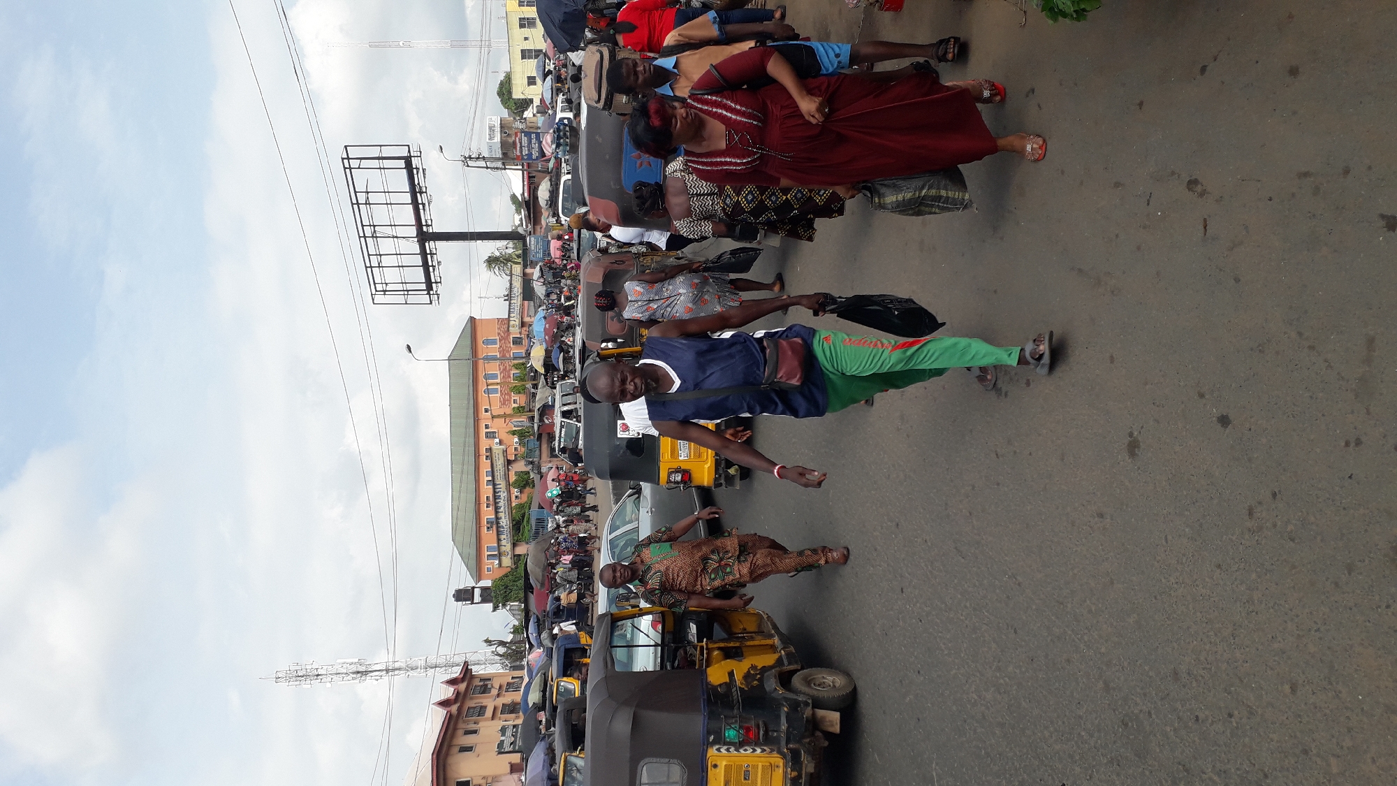 A street scene in Onitsha, Nigeria, captures a bustling intersection filled with people and vehicles. In the foreground, a man in a blue tank top and green track pants walks towards the camera, carrying a black plastic bag in his left hand and a small maroon bag across his chest. To his left, another man in a patterned short-sleeved shirt and brown pants walks in the same direction. Behind them, a yellow auto-rickshaw is parked. In the mid-ground, numerous auto-rickshaws, cars, and pedestrians are visible. People are walking, some carrying bags, and several are gathered near parked vehicles. The background features buildings with orange and brown facades, some with shops or businesses. A large billboard stands prominently in the distance, and a telecommunications tower is also visible. The sky is overcast with scattered clouds. The overall impression is that of a busy, everyday street scene in a Nigerian city.