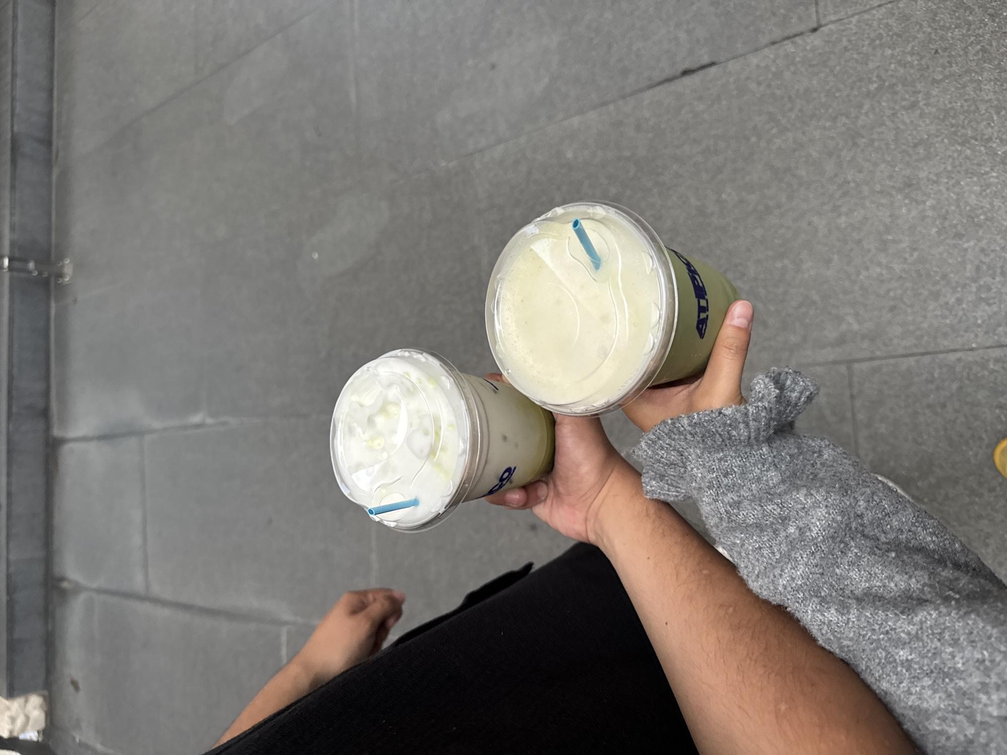 Here is a description of the image:
The image shows a person holding two beverages in clear plastic cups with lids and straws, likely matcha lattes. One latte has whipped cream on top. The person is wearing a grey knitted sweater and black pants. The setting appears to be an outdoor location with grey concrete paving. Based on the city, San Andrés Cholula, Mexico, the environment is likely a street or public space.