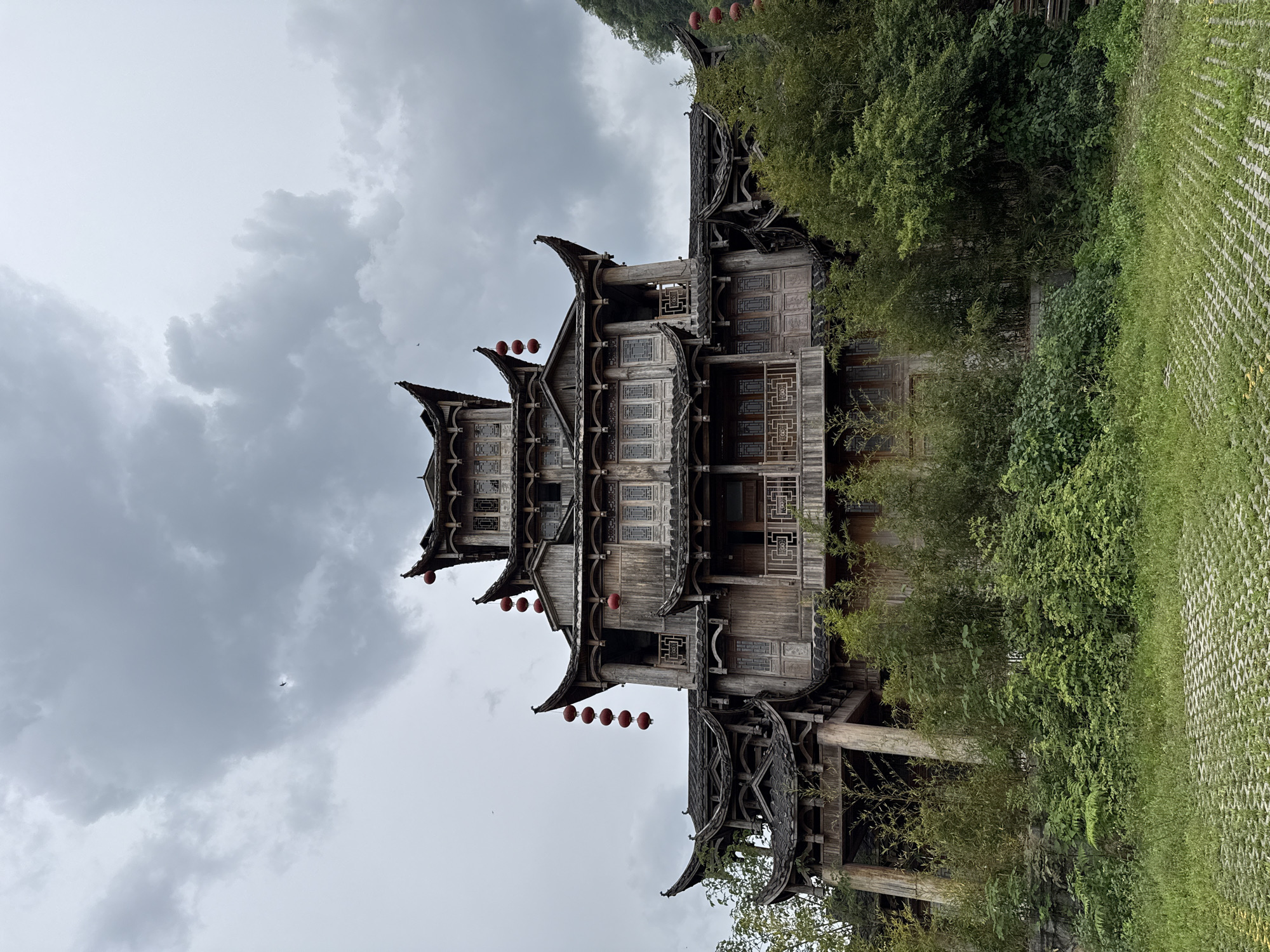 The image displays a tall, multi-story traditional Chinese wooden building with distinctive tiered, curving roofs and intricate architectural details. The structure is constructed from dark, aged wood, featuring multiple levels adorned with carved window frames and patterned latticework. Numerous red lanterns hang from the eaves at different levels, providing vibrant accents against the earthy tones of the building.
The building is nestled amidst lush green vegetation, including dense trees, bamboo, and various leafy plants that partially obscure its lower sections. To the right of the building, a patterned ground surface or path, possibly made of stone or concrete with a grid-like texture, is visible and partially covered by green groundcover.
The sky is overcast with soft, grey clouds, suggesting a cloudy or muted daylight setting. A small, dark shape, possibly an airplane, can be faintly observed high in the clouds in the upper right portion of the sky (when upright). There are no people visible, nor any discernible text. The scene evokes a sense of traditional architecture integrated with nature, indicative of a tranquil historical or cultural site within a natural landscape like Wulingyuan.