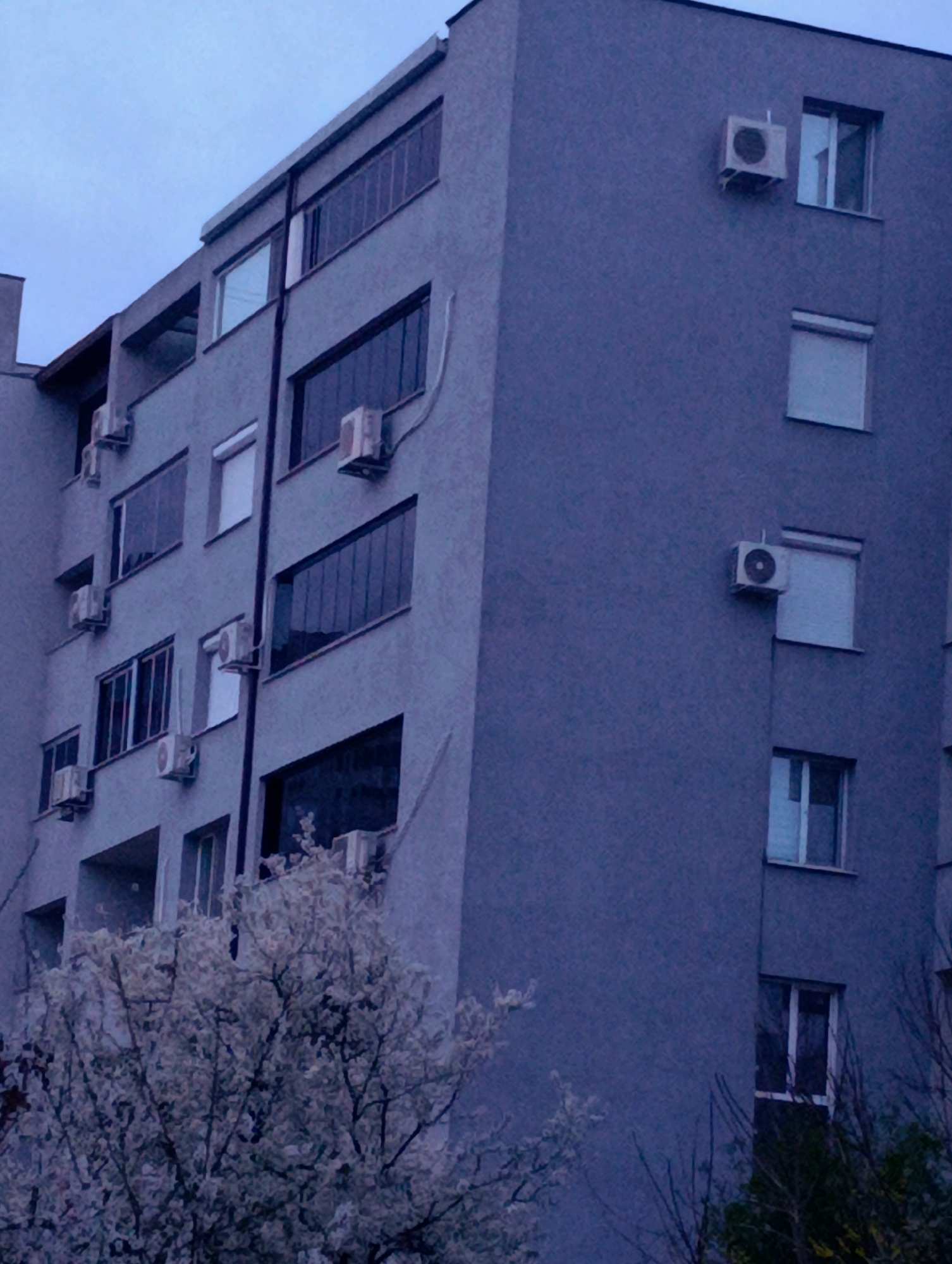 The image shows the exterior of a multi-story building with a gray facade. Several windows are visible, some with window coverings. Air conditioning units are attached to the building's exterior near some of the windows. In the foreground, there are trees with foliage, partially obscuring the lower portion of the building. Based on the presence of Cyrillic lettering on building signage, the location is likely in a Slavic country. It is specified that the location is the city of Kardzhali, Bulgaria. The sky is overcast, suggesting dusk or dawn.