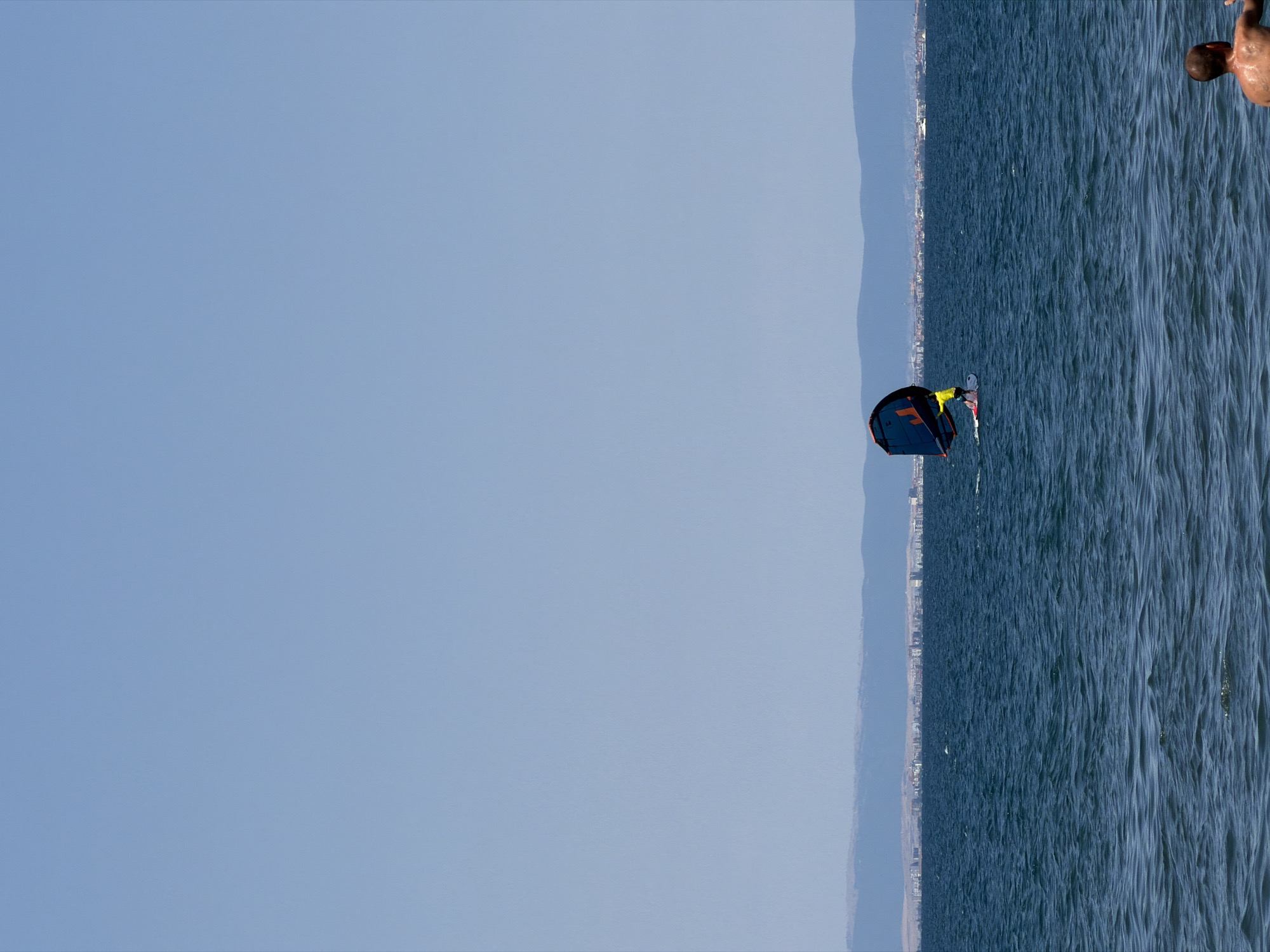 In the distance, a person is wing foiling on the Black Sea near Atiya, Bulgaria. They are wearing a yellow shirt and are on a white board, holding a blue and orange wing. The water is choppy, and a distant city skyline is visible on the horizon. In the foreground, a person is partially visible from the shoulders up, looking out at the water.