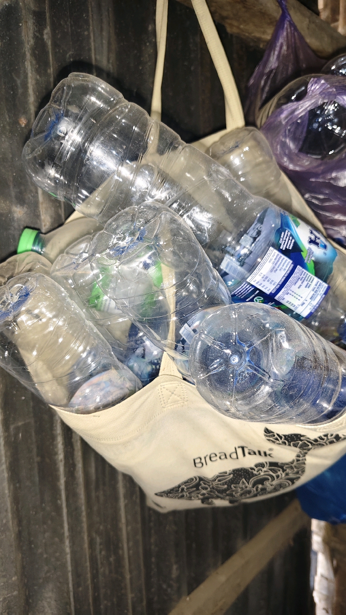 In a dimly lit, industrial or storage area, likely in Galis, Indonesia, a canvas tote bag overflowing with used plastic water bottles is prominently displayed. The bag, which features a black whale silhouette design and the text "BreadTalk" in dark lettering, is the central subject. The bottles are clear and some appear to have labels with blue and white text. The background consists of a corrugated metal wall, giving the scene a somewhat utilitarian feel. Additional items, including a purple plastic bag and other unidentifiable objects, are visible in the periphery, suggesting a location where waste or materials are being stored or collected. The lighting is uneven, creating shadows and highlighting the reflective surfaces of the plastic bottles.