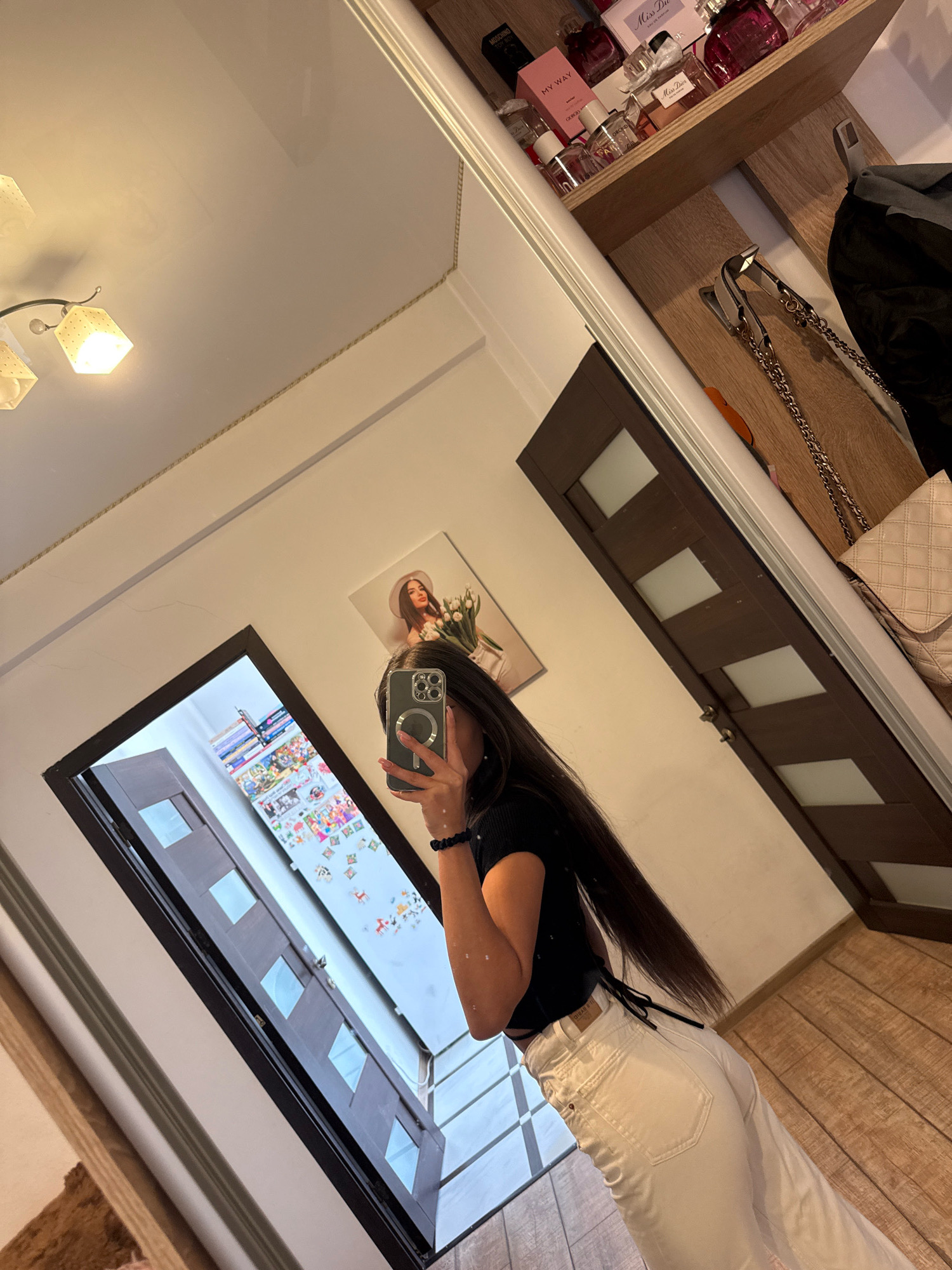 The image shows a young woman with long dark hair taking a mirror selfie in what appears to be a home interior. She is wearing a black cropped top and white wide-leg jeans. She is holding a smartphone in her right hand and her left arm is bent. In the background, a shelf is visible filled with perfume bottles. Adjacent to the shelf, a door with glass panels and a beige quilted handbag with a chain strap hanging on a hook are present. A framed picture of a woman with a hat and flowers is hanging on the wall above the door. The ceiling has decorative moldings and a light fixture with three lampshades. The floor is wooden. The overall impression is of a personal space within a residence. The location in Chișinău, Moldova, is not discernible from the visual cues in the image.