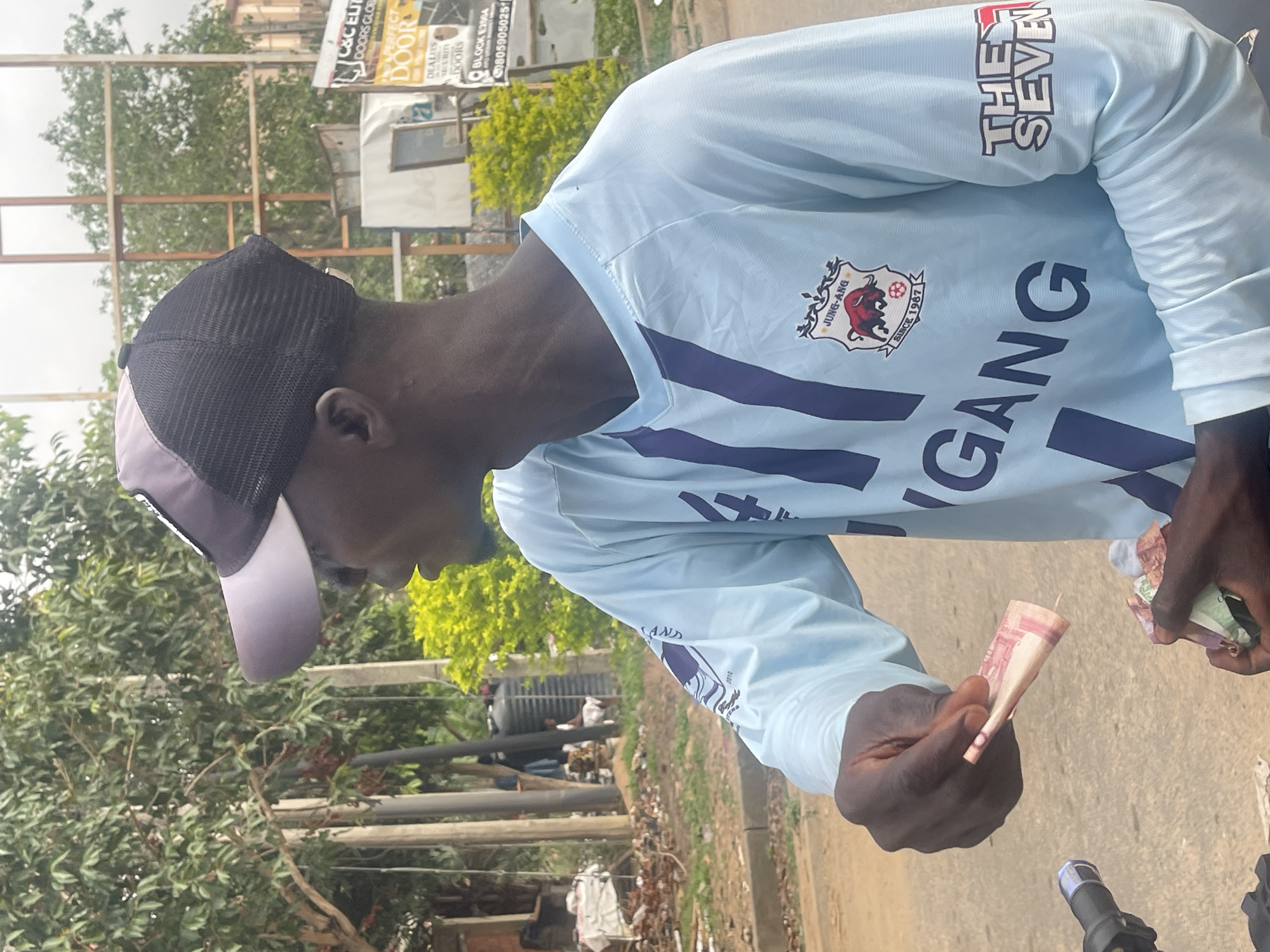The photograph depicts a man on a street in Abuja, Nigeria. He wears a light blue long-sleeved shirt with the words "Jung-Ang Gang" and a logo featuring a bull with "Since 1987" inscribed, as well as the words "The Seven" on the sleeve. He is wearing a two-tone baseball cap. He is holding Nigerian Naira notes in his hands. In the background, there are trees, vegetation, a building, and street signs, including one with "C&C Elite Doors Global" and "Perfect Door." Another sign mentions "Block E2004" and a phone number. The man is situated on a street with asphalt.