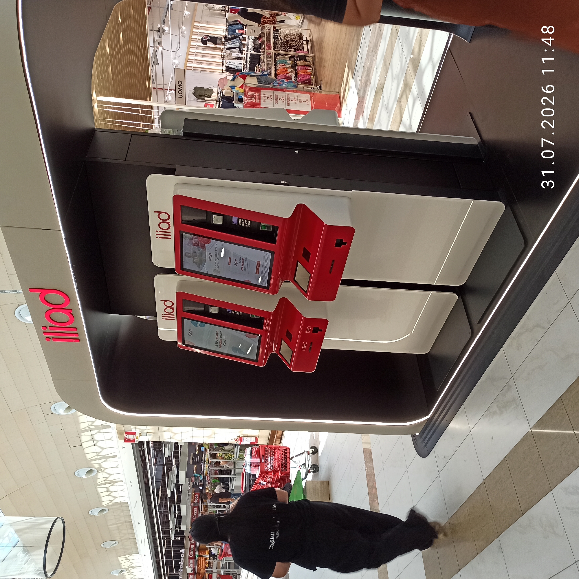 The image captures a modern retail environment, likely a shopping mall, in Modugno, Italy. In the foreground, a woman in a black t-shirt and pants walks away from the camera. She is seen from behind and her dark hair is tied back. To her right, two self-service kiosks with red frames and illuminated screens stand prominently. These kiosks appear to be branded with "iliad," a telecommunications company. The screens display graphical information, one showing text related to phone plans and the other featuring a woman.
Behind the kiosks, a clothing store is visible, with mannequins displaying outfits and racks of clothing. A sign above the store entrance reads "UOMO," indicating it is a men's clothing section. Sale signs are also visible, advertising discounts on various apparel items. In the background, other shoppers are present, and a red shopping cart filled with Coca-Cola bottles is situated near the woman in black. The floor is tiled with a pattern of light and dark beige squares. The overall lighting suggests an indoor commercial space with overhead illumination. A timestamp at the bottom right corner reads "31.07.2026 11:48."