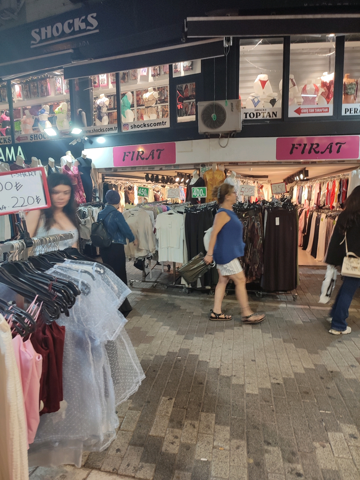 The image depicts a street-level view of a retail area, likely a market or shopping street, in Kadıköy, Turkey. In the foreground, clothing racks filled with various garments are visible, including sheer white dresses with polka dots and colorful t-shirts. A woman with dark hair is partially obscured by these racks, looking to her left. Behind her, a woman in a blue top and striped shorts is walking away from the camera, pulling a small rolling suitcase. She is wearing sandals. Further back, racks of clothing are displayed outside a shop with a pink sign that reads "FIRAT". The shop's interior, visible through a window, displays mannequins in lingerie. Above this shop, another storefront has a black sign with "SHOCKS" in large white letters. Various posters and signs, some with Turkish text like "TOPTAN" and prices in Turkish Lira (₺), are displayed on the shop fronts. The ground is paved with stone tiles. The overall impression is of a busy commercial street scene.