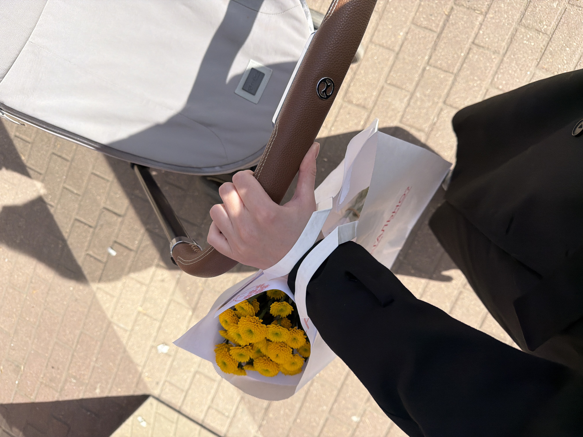 The image shows a person holding a bouquet of yellow flowers in a white paper bag, standing next to a baby stroller on a brick walkway. The person is wearing a black coat with white cuffs. The location is not specified, and there is no indication that it is in Khimki, Russia.