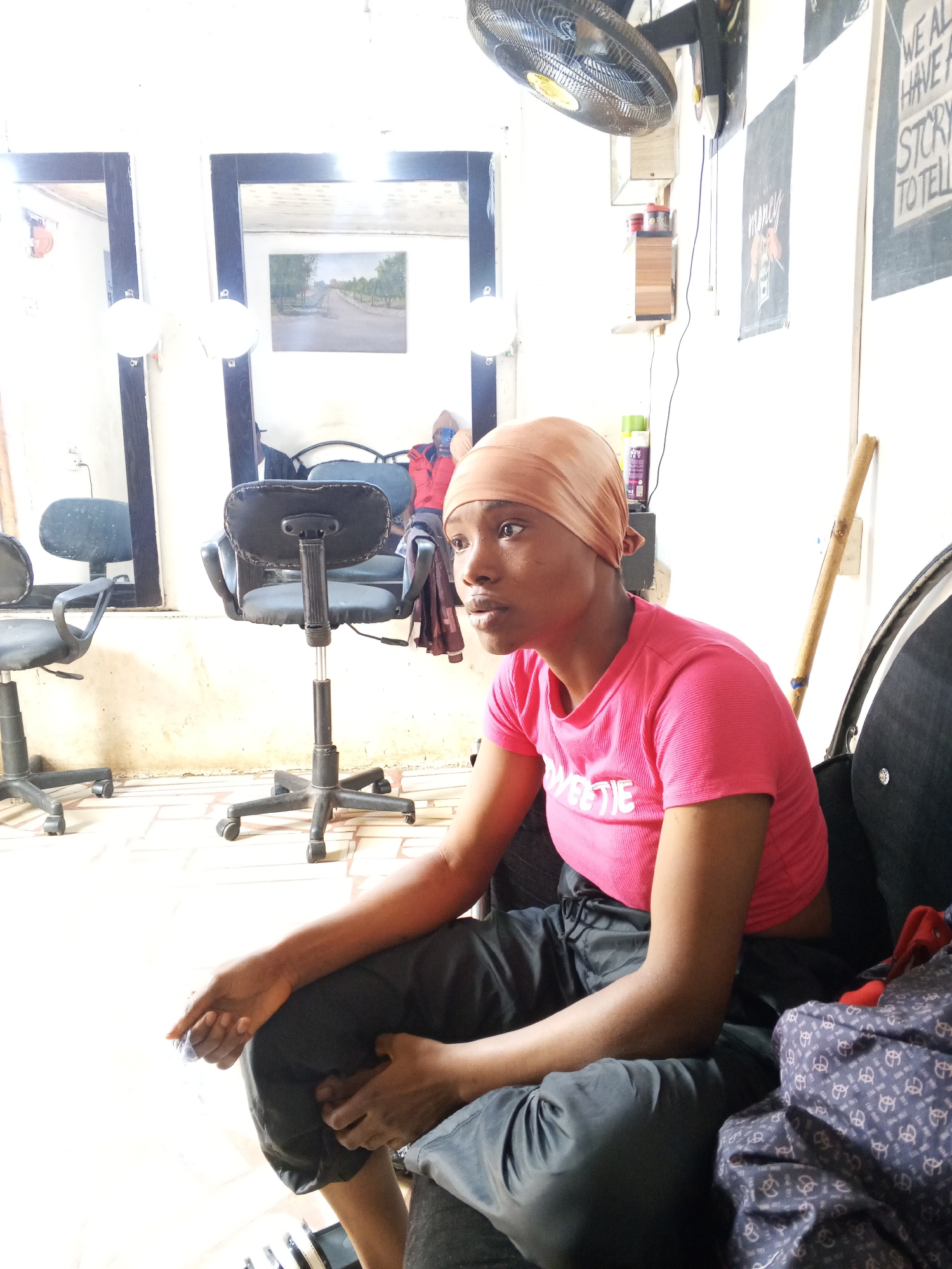 A medium shot captures a person seated in what appears to be an indoor space, possibly a hair salon, given the presence of salon chairs, mirrors with light bulbs, and hair products on shelves in the background. The location is in the city of Jos, Nigeria.
The individual has a brown head covering, wears a pink t-shirt, and dark pants. The person is seated on what appears to be a dark-colored sofa or chair. The person's gaze is directed towards the left.
The scene includes standard black salon chairs, wall mirrors with attached light bulbs, and miscellaneous items such as a standing fan, posters on the wall, and a bamboo stick leaned against the wall. The flooring appears to be tiled.