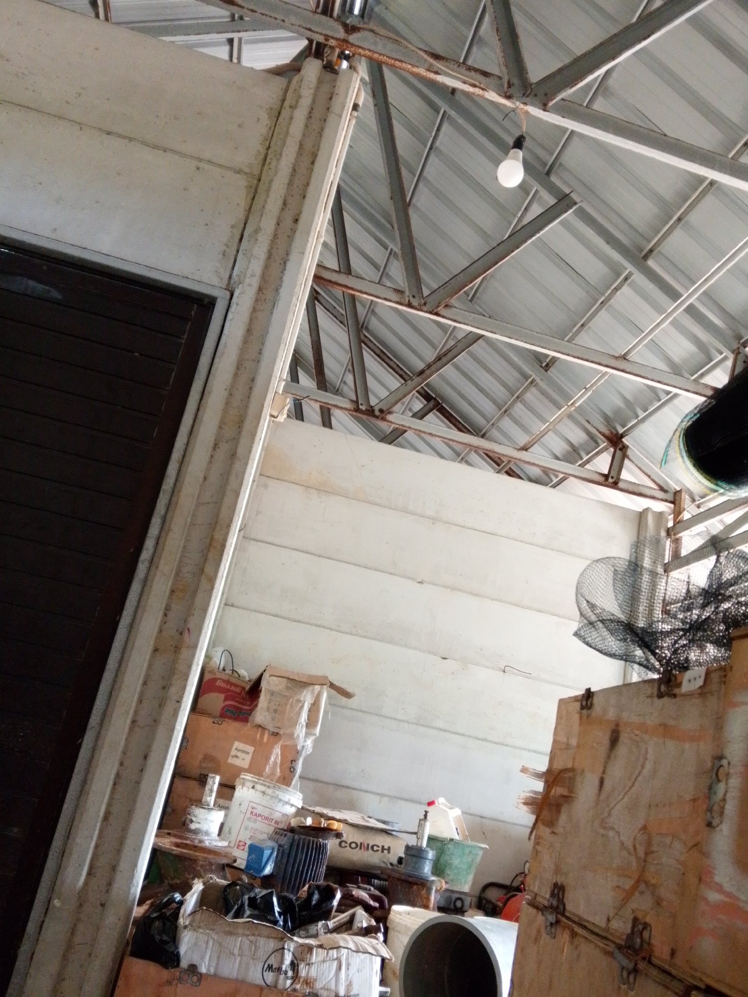 The image shows an indoor space, likely a storage area or shed, located in Bukit Ketok, Indonesia. The room is cluttered with various objects.
The structure has a metal roof supported by a steel beam framework. A single light bulb hangs from the framework. The walls appear to be made of concrete. A dark-colored door is visible on the left side.
Numerous boxes and containers are stacked on the floor and a table. A large wooden crate is on the right side, obscuring a portion of the background. Various items, including buckets, pipes, and what appears to be a net, are scattered around. The contents of the boxes and containers are partially visible, indicating a mix of household or industrial supplies.