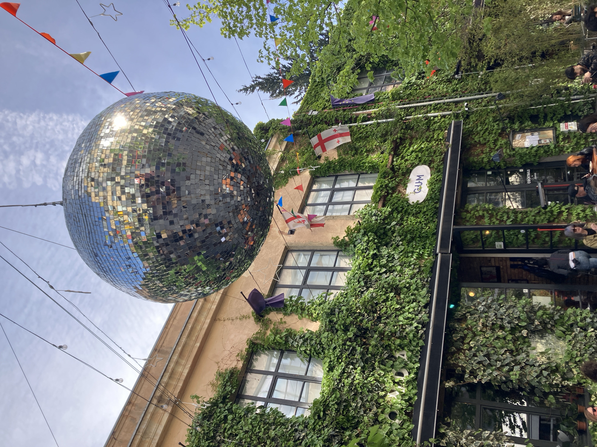 An outdoor scene in Tbilisi, Georgia shows a large disco ball suspended in the air, with colorful flags and decorative lights strung around it. A building with a beige façade is partially covered in green ivy. Windows are visible, some with black frames. Flags representing Georgia and the European Union are hanging from the building. A sign for “GUM” is visible. People are present in the background, mostly standing near the entrance of a café or establishment with the text 'cocktails coffee'.