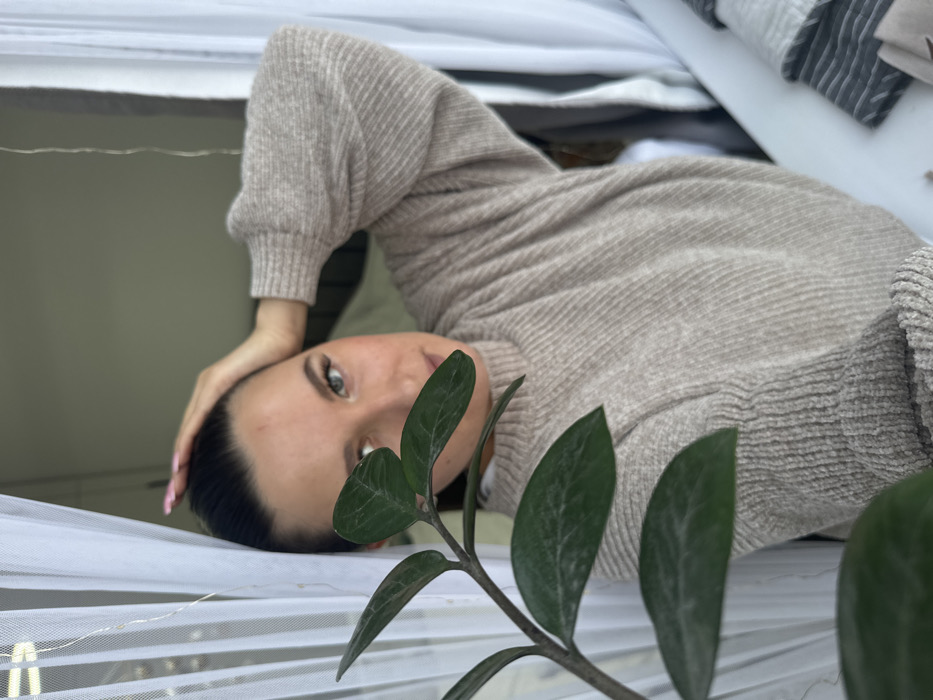 An indoor scene in Odintsovo, Russia, captures a female subject partially obscured by a dark green plant branch in the immediate foreground. The subject, positioned centrally, has dark hair pulled back and light-colored eyes, looking directly at the camera. Her right hand is raised, fingers resting on the top of her head, displaying pink nail polish. She wears a light beige ribbed knit sweater over a white undershirt. To the left, white sheer curtains are visible, interspersed with a string of small, white fairy lights. On the right side of the frame, a white surface, possibly a windowsill or table, holds folded fabrics, including one with grey and white stripes and another solid grey item. The background is softly blurred, indicating an interior room.