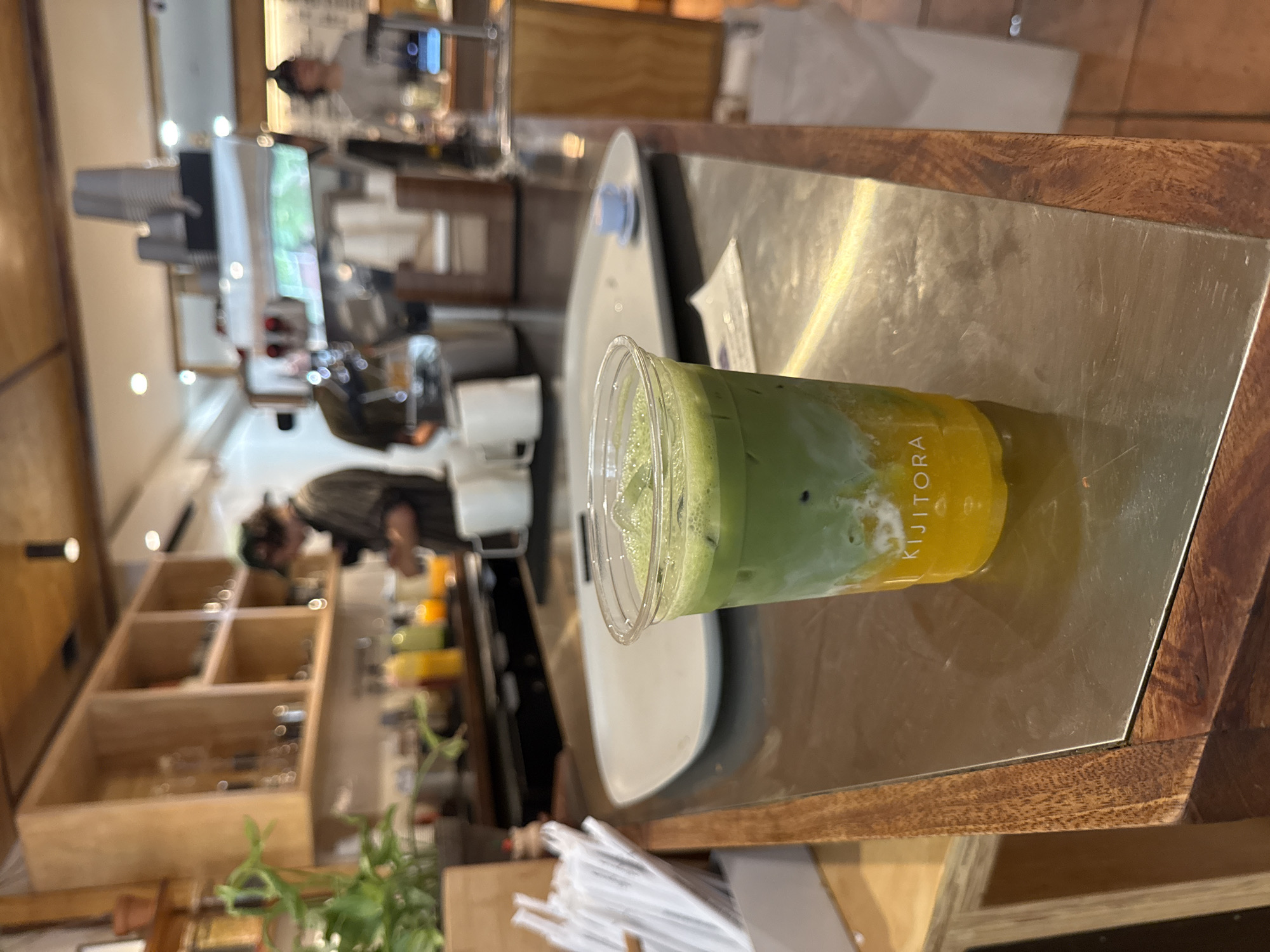 An iced matcha and mango drink sits in a clear plastic cup on a metallic counter in a cafe. The drink has distinct layers of yellow mango at the bottom, followed by a white layer, and topped with green matcha. The word "KIJITORA" is visible on the cup. In the background, shelves are stocked with various items, and people are seen working behind a counter with coffee equipment. The cafe appears to be well-lit with wooden accents. This scene is located in New York City, United States of America.