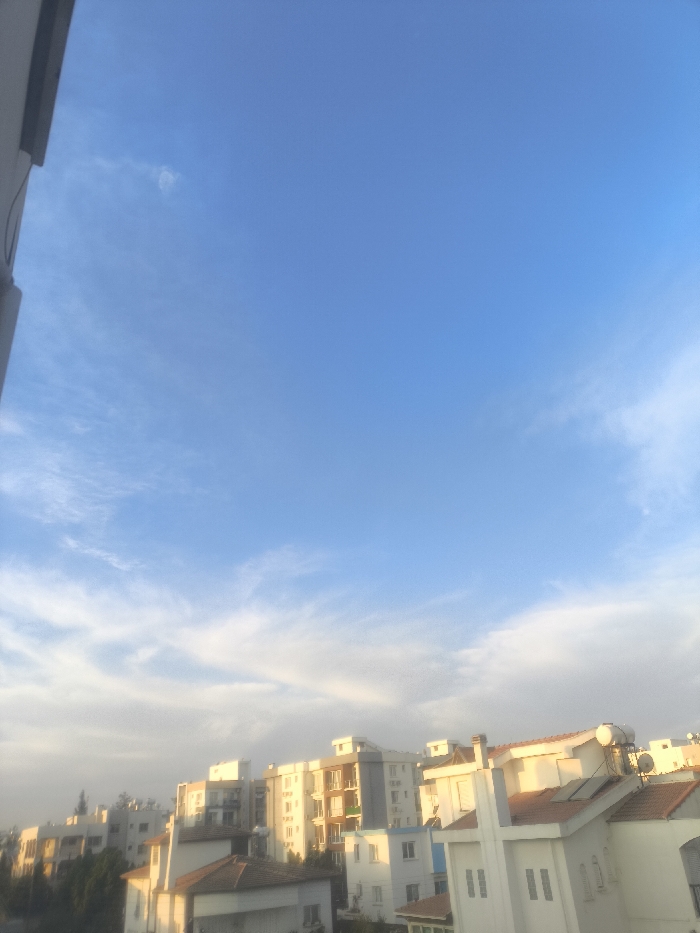 The image displays an elevated view of a residential urban area within Nicosia, Northern Cyprus / Cyprus, captured under a predominantly clear blue sky with scattered, wispy white clouds. The upper two-thirds of the frame are occupied by the sky, transitioning from a deeper blue at the top to a lighter hue near the horizon, with diffuse cloud formations. The lower third features a densely packed collection of multi-story buildings, primarily constructed with light-colored facades (white, beige, light grey). Architectural elements include flat roofs and pitched roofs with red/orange tiles. Multiple rooftops feature visible solar water heaters and photovoltaic panels. Green foliage from trees and shrubs is interspersed among the buildings, particularly on the left and center-left. A dark, vertical structural element, likely part of a building or balcony railing, frames the extreme left edge of the image. No individuals, vehicles, or discernible activities are present in the scene. The overall lighting condition suggests either early morning or late afternoon, with a warm, soft illumination on the structures.