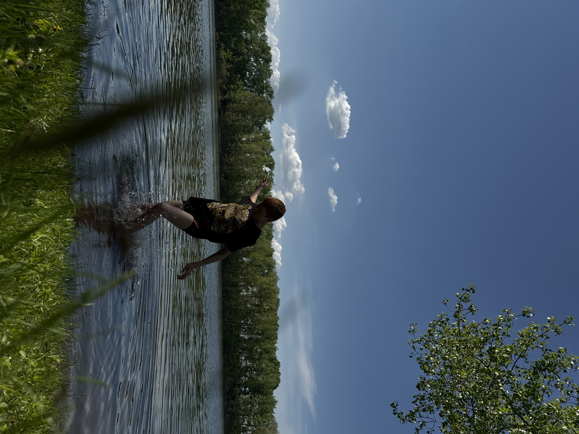 An eye-level outdoor shot of a person standing in water at a shoreline, likely a lake, in Chaikovsky, Russia. The person is wearing a dark-colored patterned t-shirt and dark shorts. The person is knee-deep in the water, with their arms outstretched to the sides, and appears to be walking into the water or has just entered the water, creating a splash. The water has ripples on its surface. The left side of the frame has a grassy shoreline. The background has a line of trees and a blue sky with some clouds. A tree branch is on the bottom right corner.