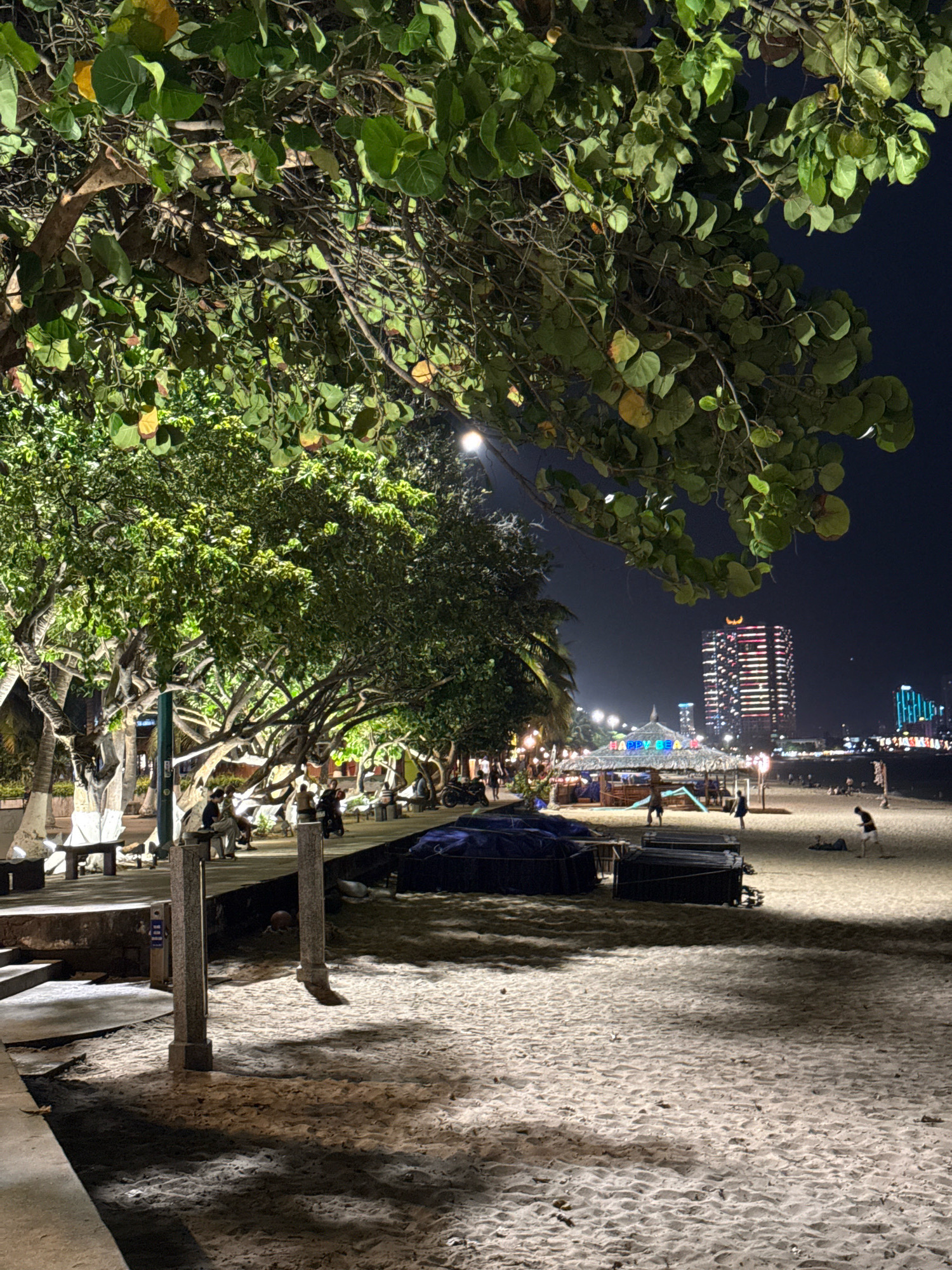 This image captures a nighttime scene on a beach in Nha Trang, Vietnam, likely along Tran Phu Beach, known for its vibrant beachfront promenade. The foreground features a sandy beach with shadows cast by overhead trees and structures. Two granite pillars stand prominently in the sandy area. Further back, a paved walkway lines the beach, with several people seated on benches beneath the canopy of large, leafy trees. The trees are illuminated by artificial lights, creating a warm glow.
Beyond the walkway, a beachside area is visible with a thatched-roof structure and some blue tarpaulin-covered objects. In the distance, the city skyline is illuminated by building lights, with a particularly tall building featuring red and white horizontal light patterns. Other structures and lights suggest a developed urban environment adjacent to the beach. Several individuals are scattered across the beach in the mid-ground and background, some appearing to be walking or engaged in casual activities. The overall impression is a lively, illuminated urban beach scene at night.