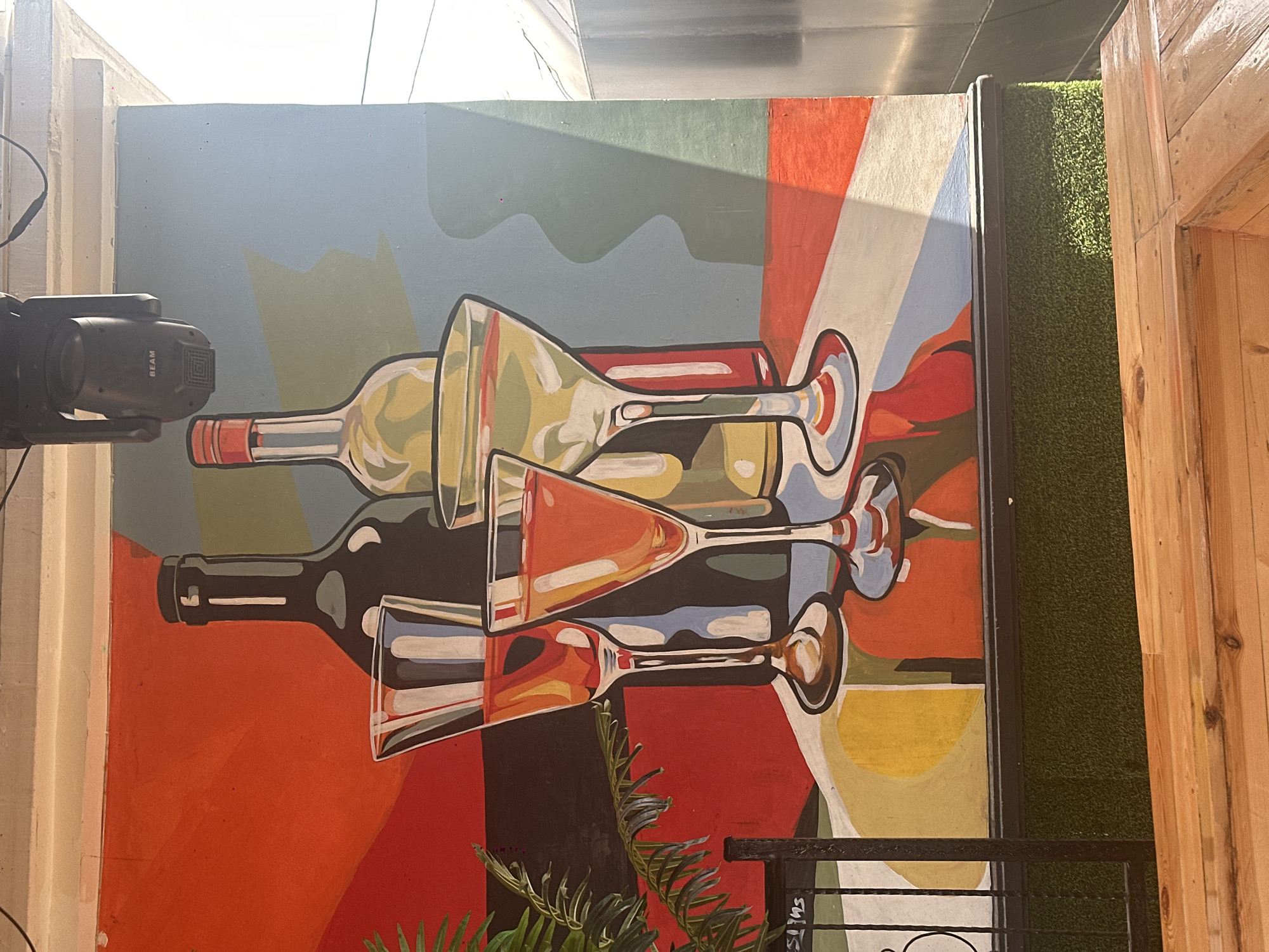 The image captures a colorful mural painted on an outdoor wall, likely in a commercial area. The mural depicts two bottles and four glasses, some filled with what appears to be red and yellow liquids. The art style is bold and graphic, with strong geometric shapes and vibrant, flat colors like red, orange, yellow, blue, and gray. The background is a mix of these colors, creating an abstract and dynamic composition.
In the foreground, there is a black metal fence and a patch of green artificial turf. Beyond the turf, a wooden structure, possibly a bench or table, is visible. The lighting suggests it is daytime, with shadows cast across the mural, indicating sunlight or artificial light sources. A black spotlight fixture is mounted on the ceiling above the mural. The overall scene suggests an outdoor seating area, possibly at a restaurant, bar, or event space. There are no people visible in the image. The location is specified as Howayi, Nigeria.