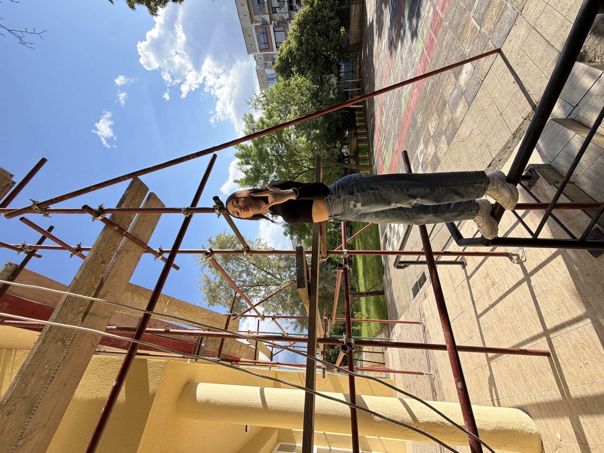 A young woman stands playfully on scaffolding in an outdoor setting, likely in Bobov Dol, Bulgaria. She is captured in a portrait orientation (rotated 90 degrees clockwise in the original image), posing with a "peace sign" or "rock on" gesture with her left hand and a pursed-lip expression. She wears a black long-sleeved crop top, ripped blue jeans, and white sneakers, with long dark hair.
The scaffolding, made of rusty red metal pipes and wooden planks, is prominently featured, surrounding a building with yellow stucco walls and a cylindrical column visible at the bottom. Wires and cables are strung across the scaffolding. In the background, green trees and parts of other buildings are visible under a bright blue sky with scattered white clouds. The ground beneath the scaffolding is paved with patterned tiles, showing distinct shadows, indicating a sunny day, likely midday or early afternoon. The overall scene suggests an informal moment captured amidst a construction or renovation area in a residential or urban environment. No visible text is legible in the image.