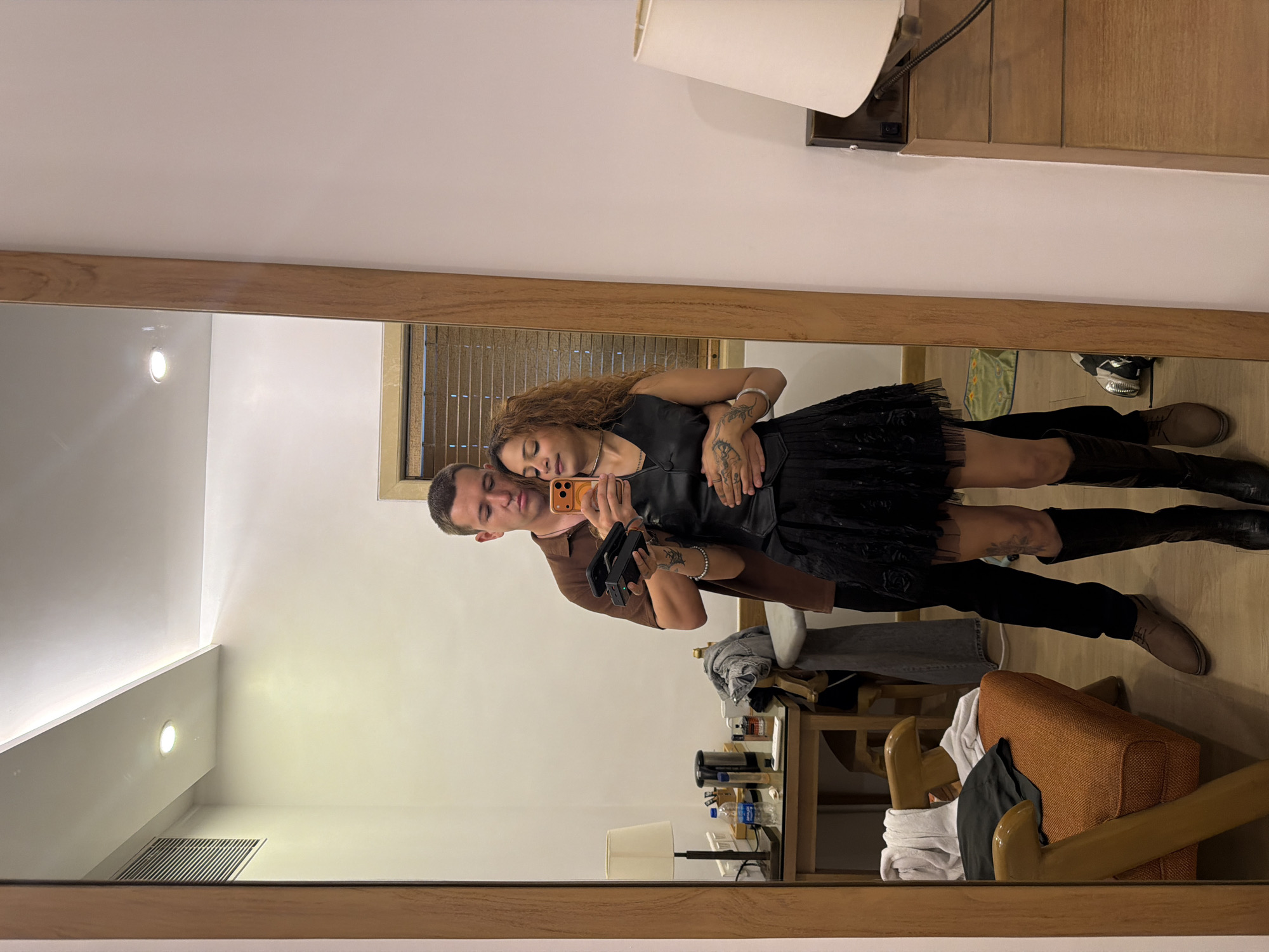 This is a full-length, indoor, eye-level mirror selfie taken in what appears to be a hotel room in Mumbai, India. A man and a woman are positioned in front of the mirror. The man, on the left, is wearing a brown short-sleeved shirt and black pants. He is holding a mobile phone in his right hand, which is raised to take the photo, and his left arm is wrapped around the woman's waist. The woman, on the right, is wearing a black sleeveless vest over a black dress, black knee-high boots, and has curly brown hair. She is holding a black object in her left hand and her right arm is resting on the man's arm. They are both looking at their reflections in the mirror.
The background shows a portion of the room. To the left, a wooden table with a lamp, some water bottles, and other items. Behind them, a window with horizontal blinds is visible. To the right, a dresser with a lamp on top. The floor is wooden. The overall lighting is soft and warm, indicating an indoor setting.