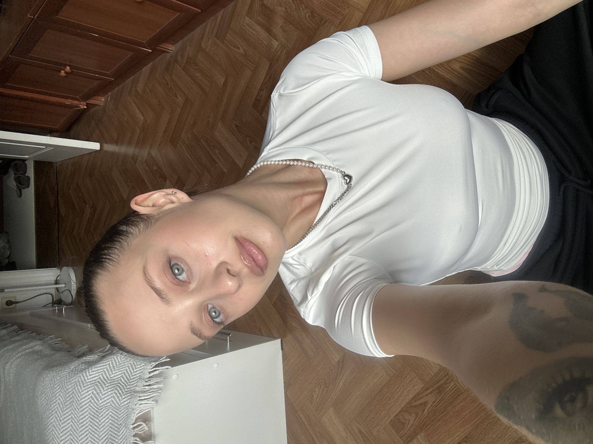 The image is a slightly angled, close-up selfie of a young woman with blue eyes and light skin. She is wearing a white t-shirt and black pants. She has a pearl necklace with a silver heart pendant. Her hair is dark and pulled back. Her face is the primary focus, and she appears to be looking directly at the camera. The background consists of a wooden floor and some furniture, including what looks like a white cabinet or dresser and darker wooden cabinets. There is also a textured blanket visible on the left side of the image. The lighting suggests it is daytime, possibly indoors. No text is visible in the image. The image appears to have been taken in Khimki, Russia.