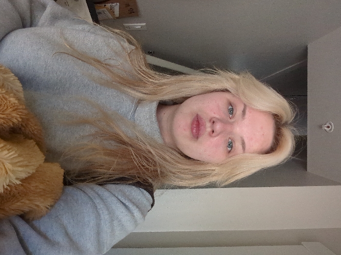 The image displays a head-and-torso view of a female individual situated indoors. The subject has shoulder-length blonde hair, light-colored eyes, and a fair complexion, facing directly towards the camera with slightly pursed lips. She is dressed in a plain light grey, high-necked, long-sleeved garment. A dark strap, possibly from a bag, is visible over her right shoulder. In the lower right foreground, a textured, light brown, furry object is partially visible. The background features plain grey walls, with a lighter grey vertical panel visible on the left and a uniformly light grey wall on the right. A light-colored ceiling with a small, circular white fixture is visible above the subject's head. There are no identifiable landmarks or environmental cues within the image to specify the location as Area D (Elaho/Garibaldi), Canada.