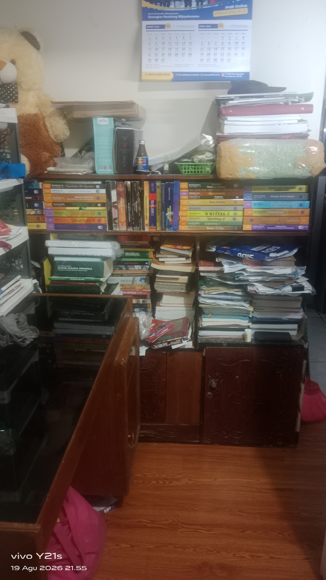 This is a full shot of a room that appears to be a study or storage area, filled with books and other items. The scene is likely indoors, possibly in a residential or office setting. The wooden floor is visible at the bottom of the image, and a wooden cabinet with carved doors is also present. The walls are likely painted a light color.
The main focus of the image is a large collection of books organized on shelves. There are many stacks of books, some neatly arranged and others piled somewhat haphazardly. Various titles and subjects are visible on the book spines. In addition to books, there are some other objects on the shelves, including what appears to be a teddy bear, a bottle, a basket, and some packaged items. The overall impression is one of a densely packed environment with a significant amount of reading material.
The image also contains a calendar on the wall, which is partially visible. The calendar shows dates for March and April, with some text in Indonesian. This suggests the location is in Indonesia, and specifically the provided crops identify the location as "Kober, Indonesia". The text "vivo Y21s 19 Agu 2026 21.55" is overlaid on the bottom of the image, indicating the model of the phone used to take the picture and the date and time it was taken.
There are no people visible in the image. The actions or interactions are limited to the arrangement of objects. The environment appears to be indoors, with artificial lighting. The overall state of the room suggests a utilitarian space dedicated to storing books and possibly other personal belongings.