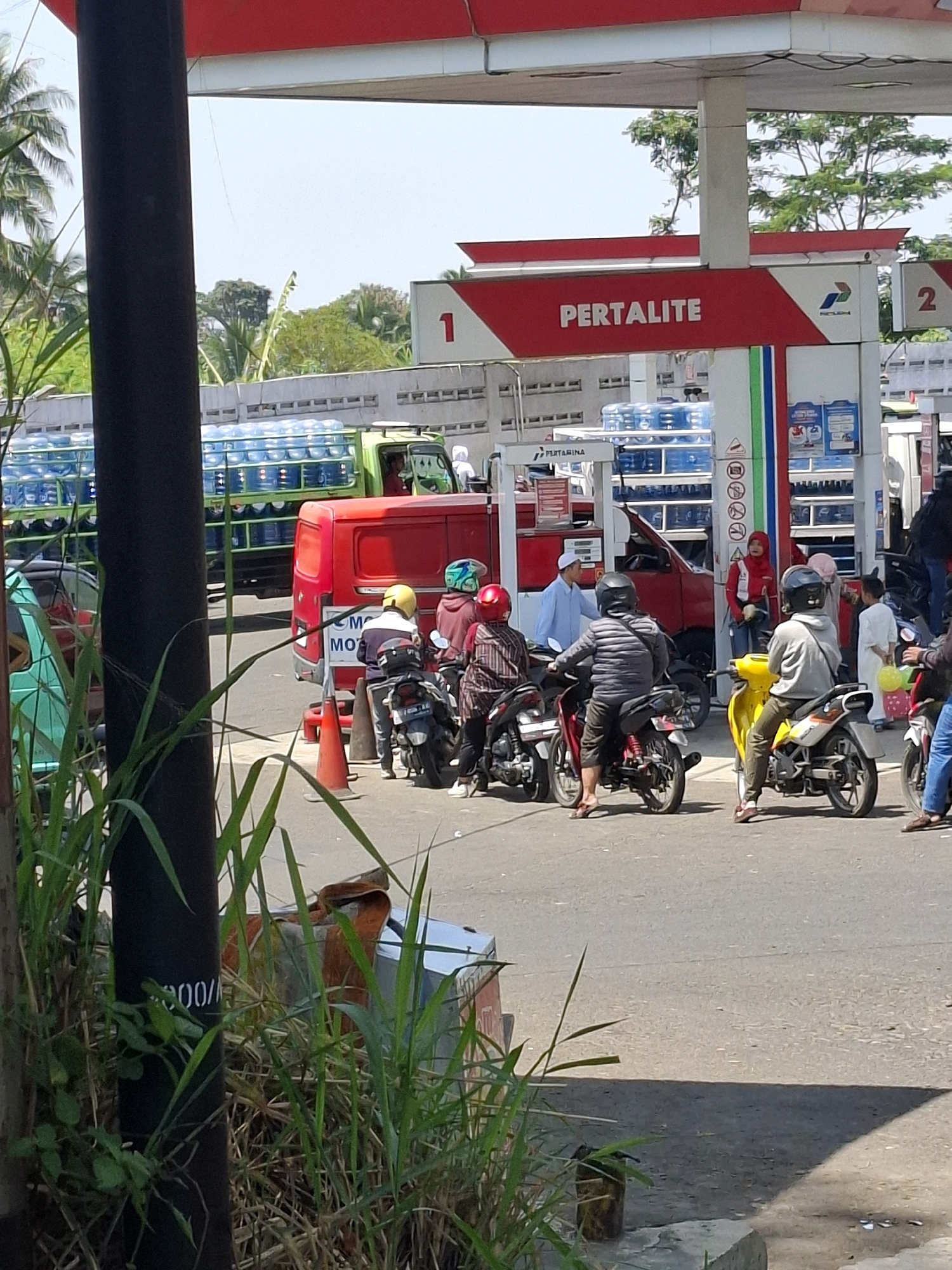 The image depicts a busy scene at a Pertamina gas station in Cikahuripan, Indonesia. Several motorcycles are lined up, with riders waiting. One motorcycle in the foreground is yellow, and another is red. A red truck is parked behind the motorcycles, and a green truck loaded with blue water jugs is also visible. A person in a red jacket and headscarf stands near the gas pumps. Tall grass and foliage are present in the foreground, partially obscuring a black pole with white lettering. The sky is clear and blue, and the overall environment appears to be sunny.