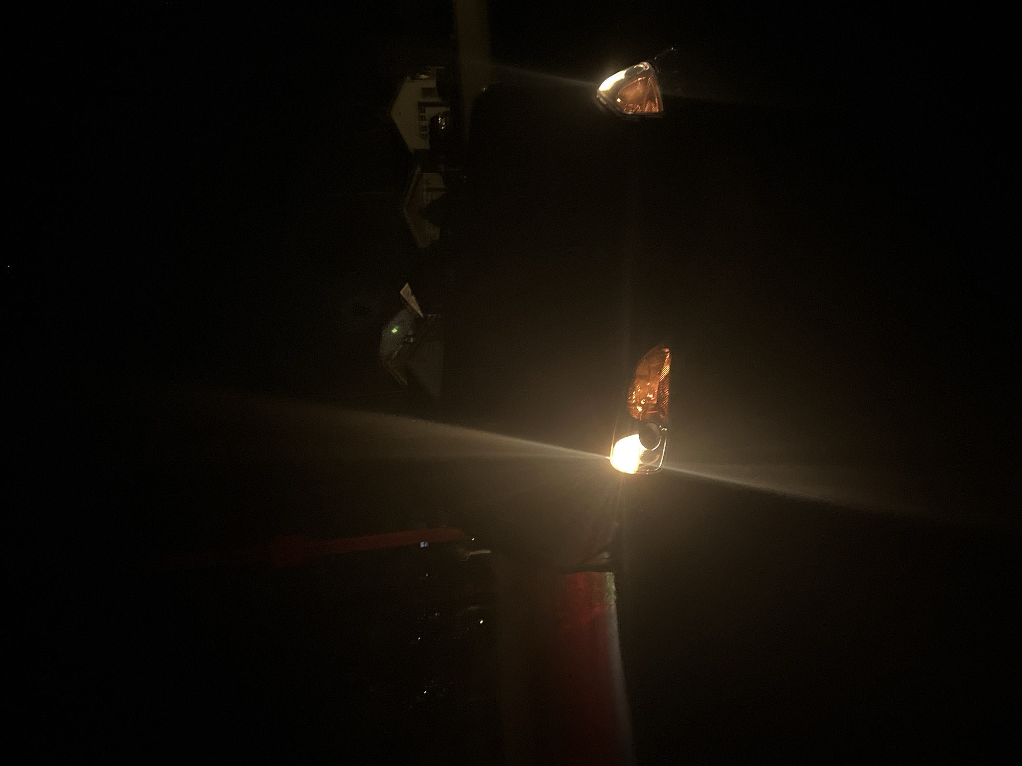 At night in the City of Newburgh, United States of America, a traffic light illuminates the darkness. The light, mounted on a pole, features amber lenses. The scene is mostly dark, with the light providing the primary source of illumination. A blurred object, possibly a vehicle, is visible in the lower left.