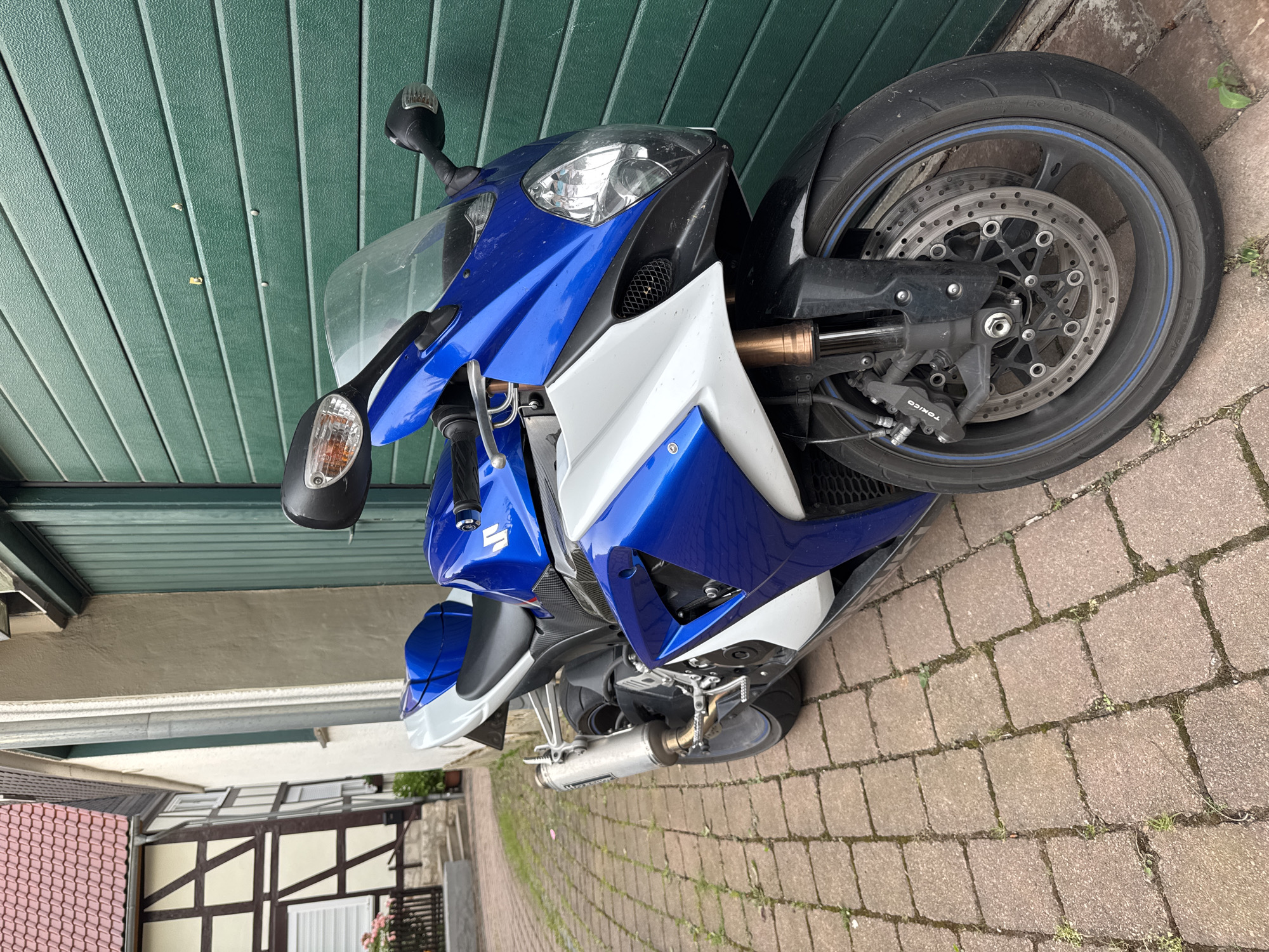 A blue and white Suzuki motorcycle is parked on a cobblestone path in front of a closed green garage door. To the left of the motorcycle, a section of a half-timbered house with a red tile roof is visible. The scene appears to be in a residential area.