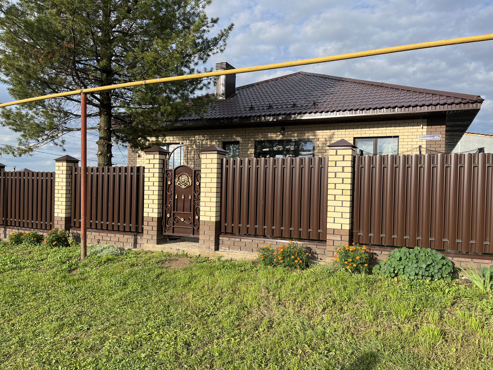 The image captures a residential property in Ufa, Russia, as indicated by a street sign reading "Деревянная Водоraz 45А" which translates to "Wooden Vodraz 45A." The scene is dominated by a single-story house constructed with light-colored brick and a dark brown, corrugated metal roof. A prominent feature is the tall, dark brown metal picket fence that encloses the property, with decorative pillars made of alternating light brick and dark stone. An ornate, dark brown metal gate is situated within the fence.
In the foreground, a lush green lawn covers the ground, with scattered patches of small orange flowers near the fence. To the left, a mature evergreen tree stands tall, its branches extending over the fence. A yellow horizontal pipe, possibly a gas line, runs across the upper portion of the image, supported by a red metal pole. The sky is partly cloudy, with patches of blue visible. The overall impression is that of a well-maintained suburban home.